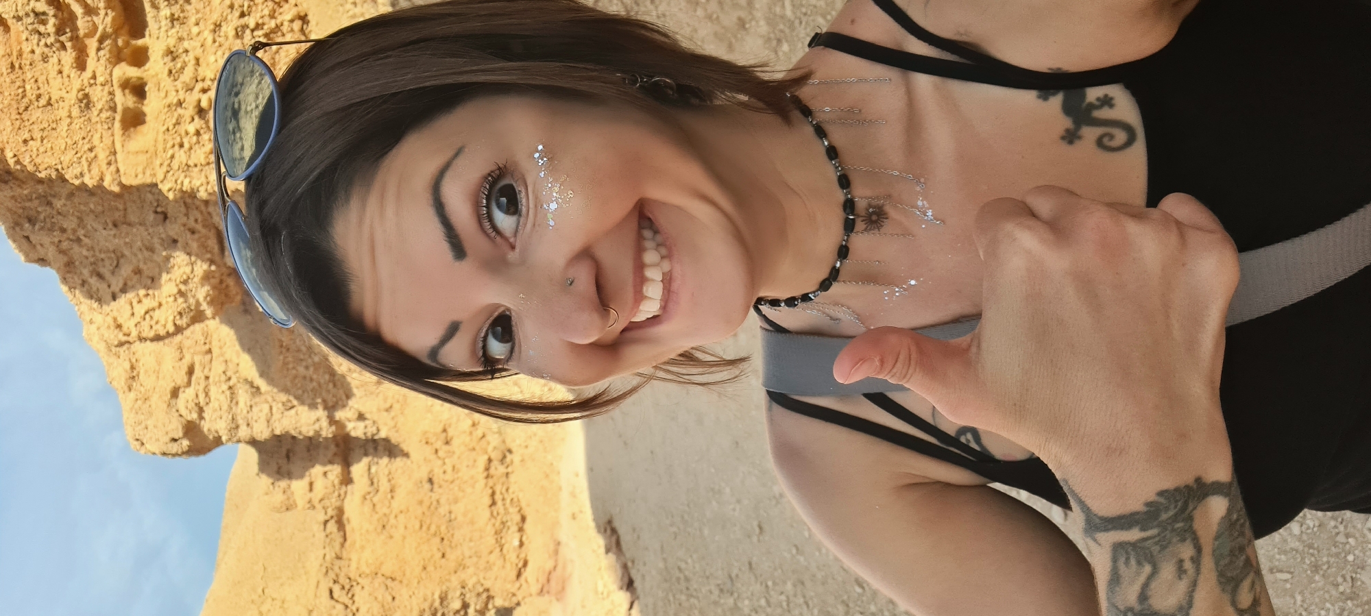 The image shows a young woman with shoulder-length dark hair. She is smiling broadly and giving a thumbs-up. She has light skin and is wearing makeup, including silver glitter near her eyes. She has a nose piercing. Her sunglasses are resting on her head. She is wearing a black tank top with spaghetti straps and a black beaded necklace with silver chains hanging from it. She has tattoos on her collarbone and wrist. She is standing outside near a tan-colored rock formation or wall, with blue sky visible in the background.
Given the presence of the tan-colored rock formation and the sky in the background, it is difficult to determine a more precise location within the Hevel Eilot Regional Council area without additional contextual cues in the photograph.