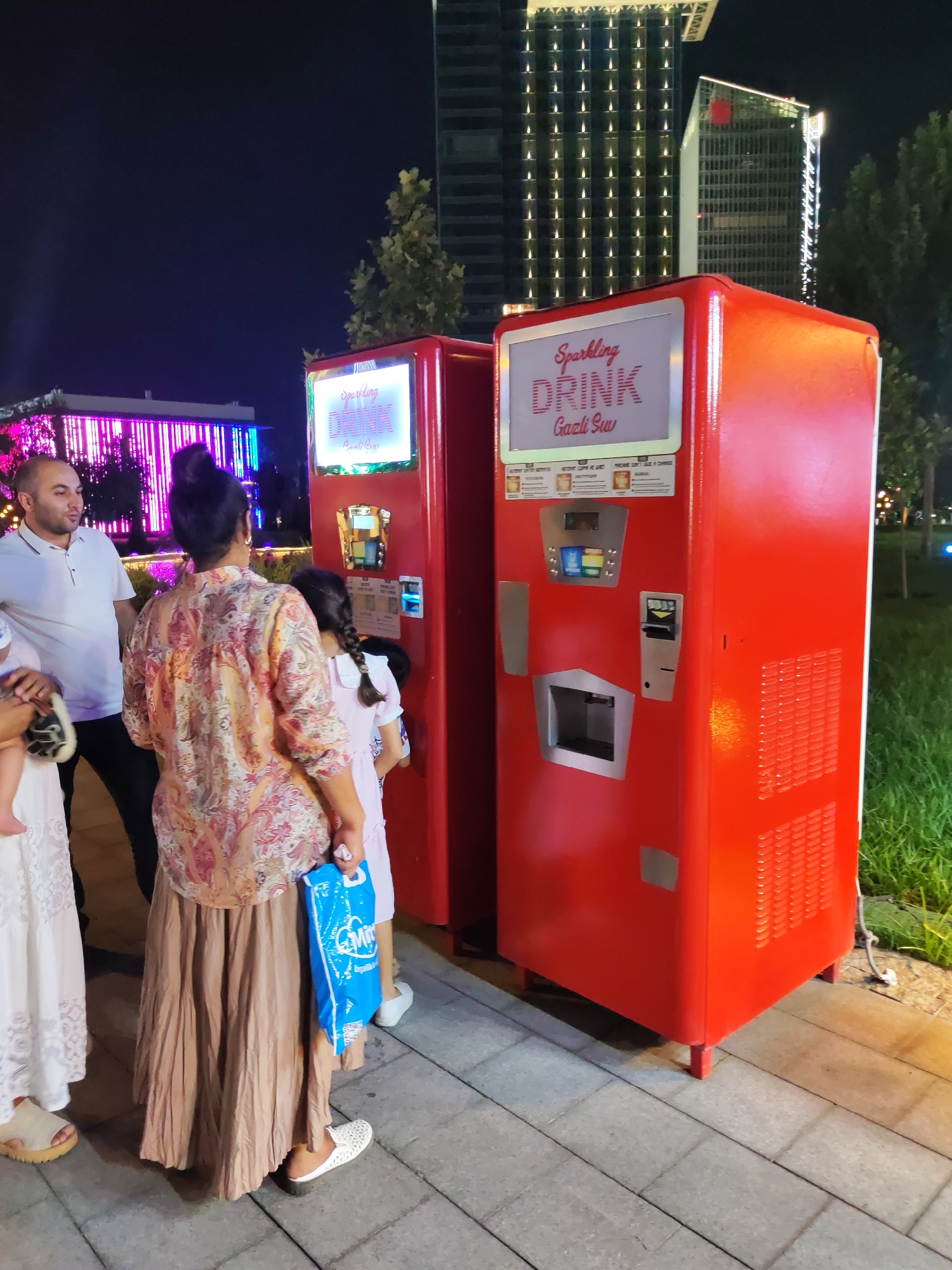 In Tashkent, Uzbekistan, at night, two red beverage vending machines stand side-by-side on a paved pathway. The machine on the right is slightly larger and has a digital display screen at the top showing "Sparkling DRINK Gazli Suv" in stylized font, along with other text and graphics indicating drink options. Below the screen, a payment and selection interface is visible, including a coin slot, card reader, and buttons. To the left of this machine is a smaller red machine with a similar digital display.
Several people are interacting with the vending machines. A man in a white polo shirt is standing to the left, looking towards the machines. A woman in a patterned shirt and a long, flowing tan skirt is standing in the center, holding a blue bag with a white logo. A young girl with braided hair, wearing a light pink dress, is standing in front of the right machine, appearing to be making a selection. Another person, partially visible, is holding a baby on the far left.
The environment suggests an outdoor urban setting at dusk or night. In the background, tall modern buildings with illuminated windows are visible against a dark sky. To the right of the vending machines, lush green grass and trees are present. The lighting is a mix of ambient city lights and the illumination from the vending machines and nearby buildings, creating a vibrant atmosphere.