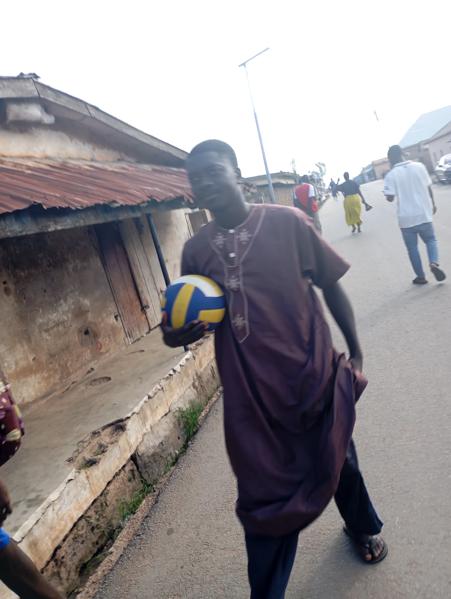 A young man wearing a purple robe and dark pants holds a volleyball in his left hand as he walks on a paved road in Shendam, Nigeria. The volleyball is blue, yellow, and white. The man is looking to his right. In the background, other people are walking on the road. To the left of the road, there is a building with a rusty corrugated metal roof and wooden doors. A concrete drainage ditch runs along the side of the road. The sky is bright and overcast.