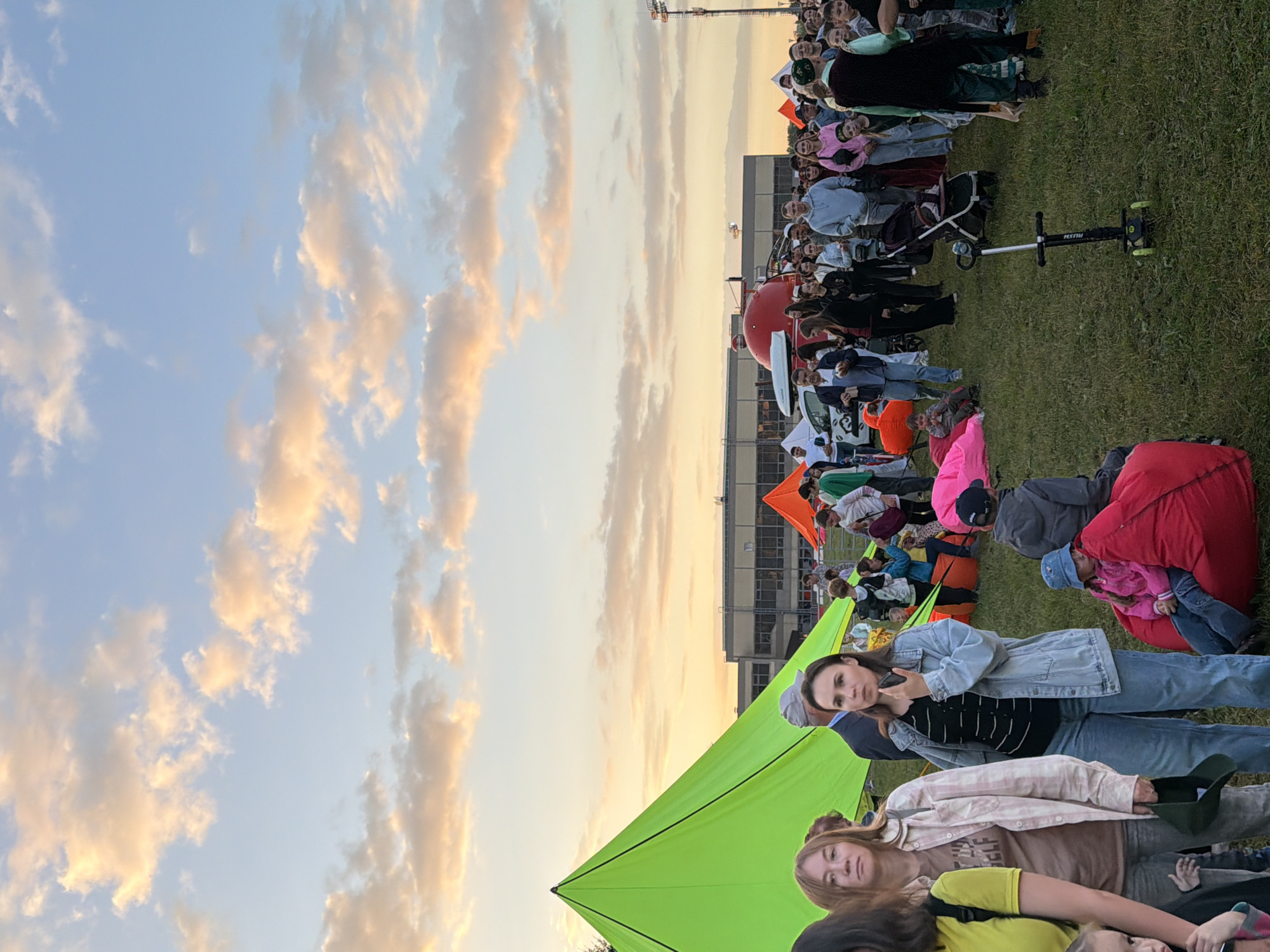 This media file depicts a gathering of people outdoors, likely at dusk. The sky is visible, filled with soft, wispy clouds illuminated by the warm tones of sunset. A large, bright green tarp is a prominent feature, creating a canopy over some of the individuals. Scattered around the scene are various objects, including beanbag chairs in red and pink, a stroller, and a kick scooter. A red, rounded structure, possibly a large inflatable or a piece of equipment, is also visible in the background.
The people appear to be seated and standing on a grassy area, facing a direction that suggests they are watching something or participating in an event. Their expressions and postures suggest a relaxed, casual atmosphere. Some individuals are looking towards the camera, while others are engaged with their surroundings or fellow attendees. The time of day is indicated by the sunset lighting, casting a warm glow over the scene. The weather appears to be clear and pleasant.
The overall impression is of an outdoor event, possibly a concert, movie screening, or community gathering, taking place in a park or open field. The presence of a building in the background might suggest an urban or suburban setting, though the exact location remains unknown. The image captures a moment of shared experience and communal enjoyment under a beautiful evening sky. No discernible text is visible in the media file.
