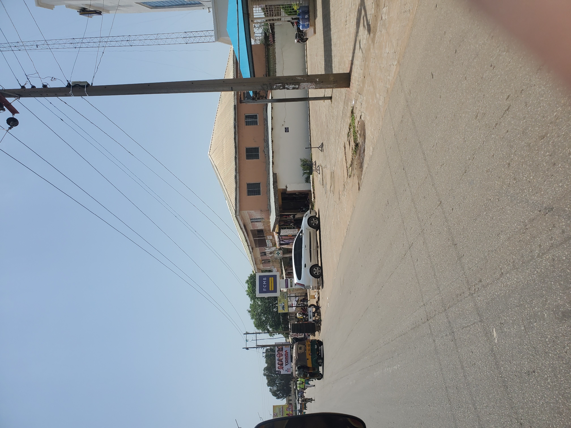 The image depicts a street scene in Jos, Nigeria, under a clear, bright sky. The foreground shows a paved road with tire marks. To the right, a concrete sidewalk is visible, with a large utility pole and overhead power lines crossing the scene. Beyond the sidewalk are buildings. One building is a two-story structure with a light-colored, sloped roof and pale orange walls, featuring several windows. Next to it, a modern white building with a blue canopy extends over an open area. Parked in front of this building are several large bottles of water. A motorcycle is also parked nearby.
On the left side of the road, a busy street is in view. Several vehicles are present, including a white SUV parked on the side of the road, and a colorful auto-rickshaw (keke napep) with passengers. A few pedestrians are walking along the side of the road. Billboards and signs are visible, including one for "FCMB". The overall impression is of a typical urban environment in Jos, with ongoing commercial activity and daily life.