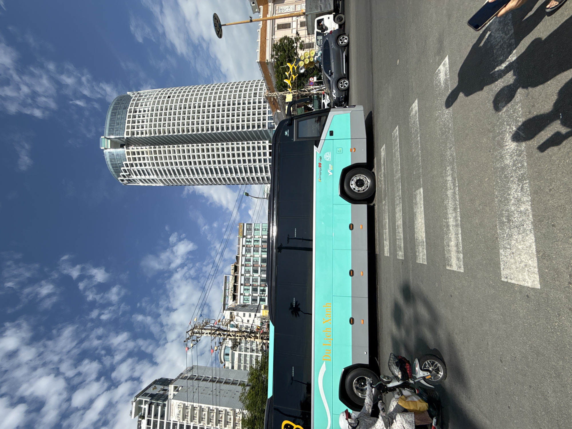 A wide-angle, eye-level shot captures a street scene in Nha Trang, Vietnam. In the foreground, a pedestrian crossing with white stripes is visible on a paved road. A teal and grey bus with tinted windows is driving across the crossing, and a person on a scooter wearing a white helmet and patterned clothing is riding in front of the bus on the left side of the frame. In the midground, several cars, including a dark grey SUV and a white truck, are stopped at the crossing or driving behind the bus. Behind the vehicles, a row of buildings of varying heights and architectural styles lines the street, with a tall, cylindrical white skyscraper with a glass-topped roof dominating the background. The sky above is a clear blue with scattered white clouds. The image appears to have been taken on a sunny day.