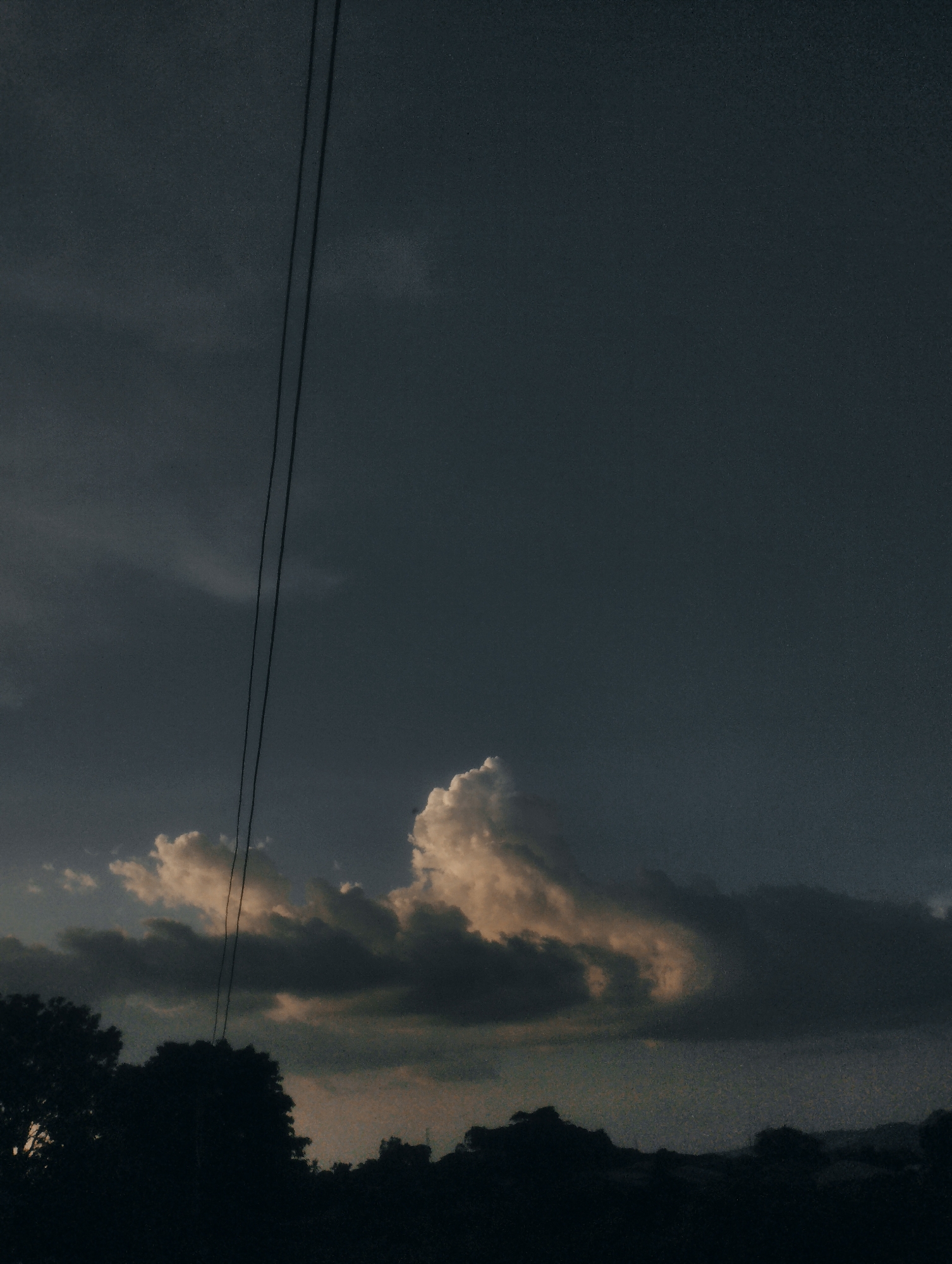The image depicts a dusky sky over a silhouetted landscape. Two dark power lines stretch diagonally across the frame from the upper left towards the center. Below the power lines, a dramatic cloud formation is visible. The clouds are a mix of dark grays and lighter, illuminated whites and oranges, suggesting the last rays of sunlight breaking through or reflecting off them. The bottom of the image features a dark silhouette of trees and possibly a low horizon line, indicating a rural or semi-rural setting. The overall mood is somber and perhaps a bit dramatic due to the lighting and cloud cover. The time of day appears to be late evening or early morning, just before sunrise or after sunset, given the low light and the warm tones in some of the clouds. There is no visible text in the image. The setting, with the power lines and silhouetted trees, is consistent with a location in Jos, Nigeria, during twilight.