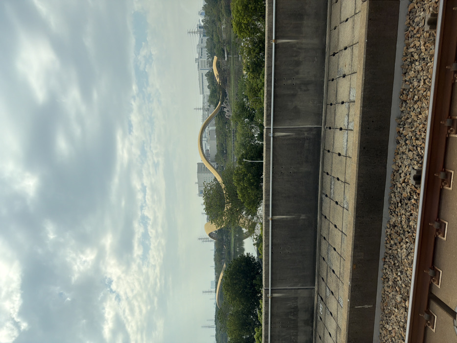 An eye-level shot of a wavy golden architectural structure across a river with trees, as seen from an elevated position. A concrete wall and railroad tracks are to the right of the frame. The sky is mostly cloudy, obscuring the buildings in the background. The scene is bathed in a muted, diffused light.