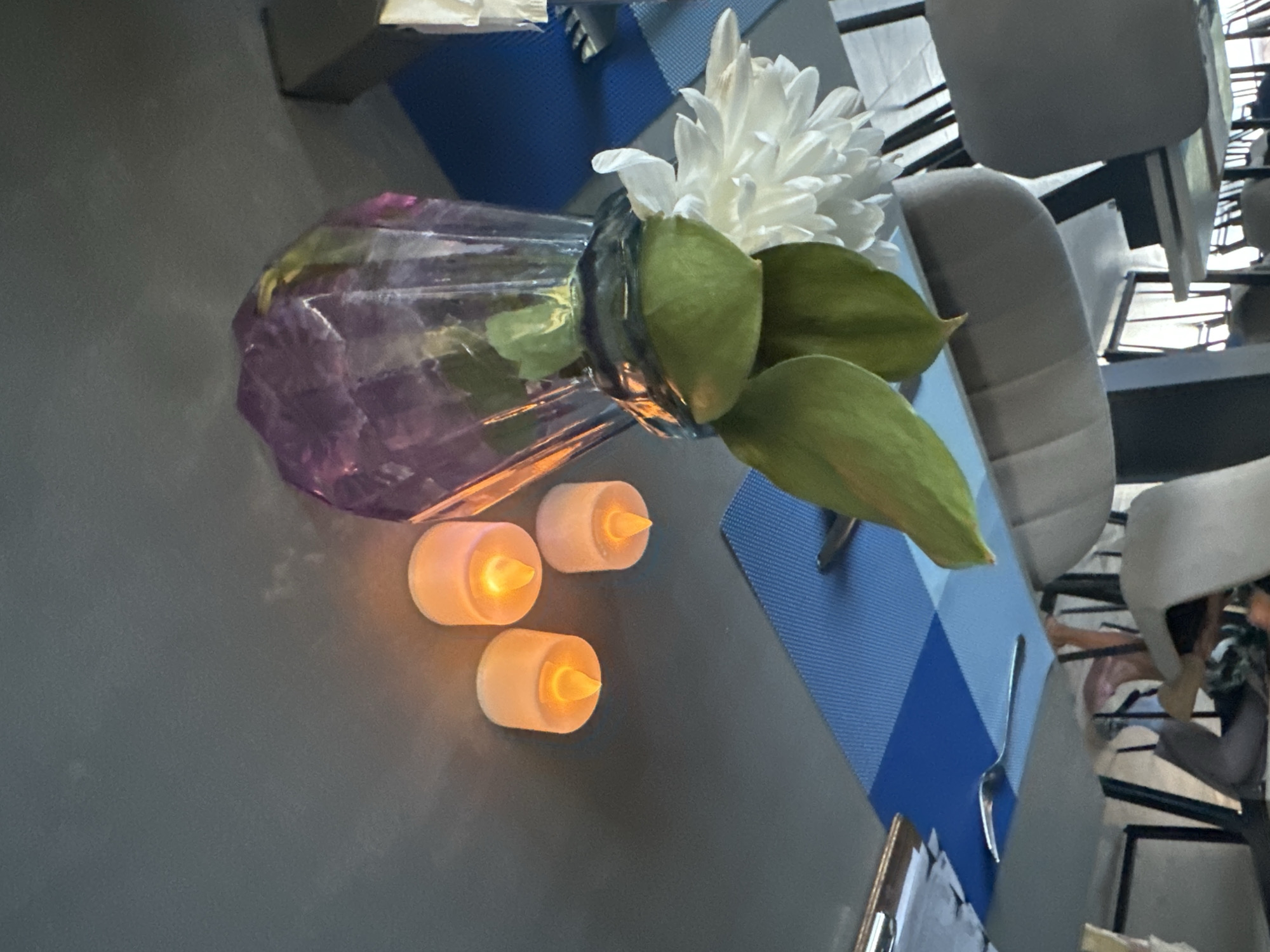 The image depicts a table setting in what appears to be a restaurant. In the foreground, a purple glass vase holds a white chrysanthemum and some green foliage. Three small, battery-operated candles with orange flames are arranged to the right of the vase. The table is covered with a gray surface, and a blue checkered placemat is visible behind the vase. In the background, several chairs and other tables are present, suggesting an indoor dining area. The overall scene is well-lit, with the candles providing a warm glow. There is no visible indication of the specific location being in Даниловка (алтайский), Russia.