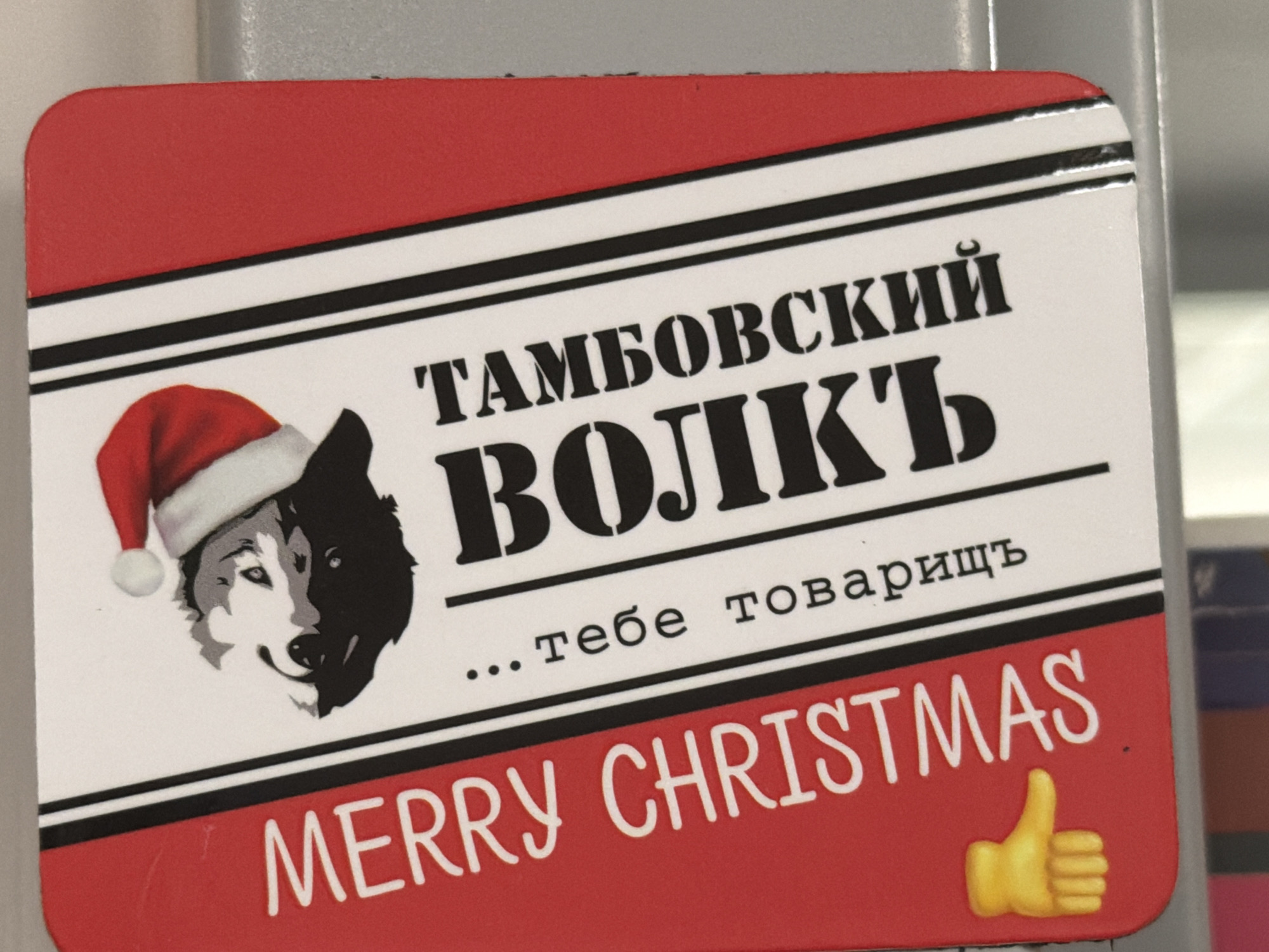 A rectangular sticker with rounded corners features a red border at the top and bottom, separated by white stripes with black lines. On the left side of the white area, a wolf wearing a Santa hat is depicted, with its face split between black and grey. The text "ТАМБОВСКИЙ ВОЛК" is prominently displayed in black, stencil-like font. Below this, in a smaller font, the Russian phrase "... тебе товарищ" (which translates to "...to you, comrade") is written. The bottom red section contains the English text "MERRY CHRISTMAS" in white, embossed lettering, accompanied by a yellow thumbs-up emoji on the right side. The sticker appears to be affixed to a light grey, metallic surface, possibly a refrigerator or a locker. There are no people or animals present in the immediate scene other than the depicted wolf. The environment appears to be indoors, likely a domestic or office setting. No specific location in Framingham, Massachusetts, USA can be identified from the image.