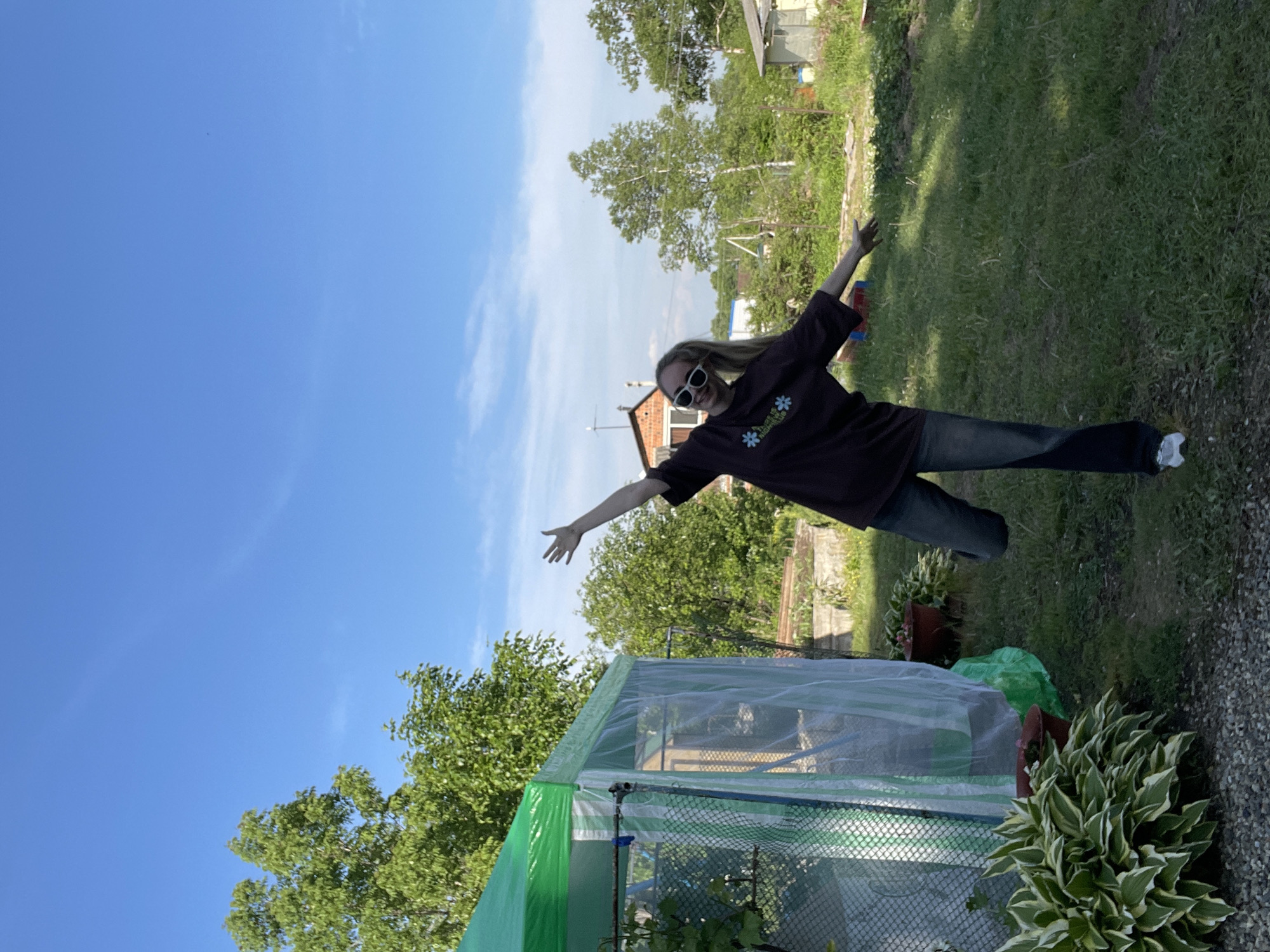 A woman stands in a grassy yard in Yasnoe, Russia, arms outstretched as if in flight. She wears sunglasses, a brown shirt with floral designs, and jeans. Behind her is a clear blue sky with a few clouds. A greenhouse with green trim is visible in the foreground. In the background are trees and a building. The yard also contains greenery and potted plants.