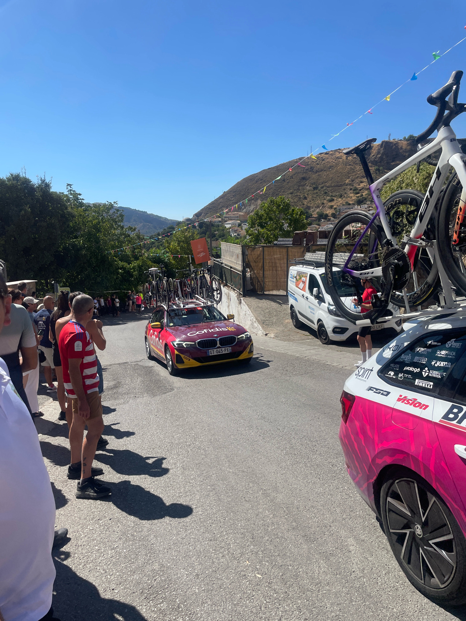 The image depicts a street scene in Güéjar Sierra, Spain, on a clear, sunny day. A red car with "Cofidis" branding and bicycles on its roof is driving down a paved road. A white van is parked to the right of the car. Several people are standing on the left side of the road, watching the car pass. A pink and white car with various sponsor logos is parked in the foreground on the right. The background features a hillside with trees and buildings, and colorful triangular flags are strung across the street.