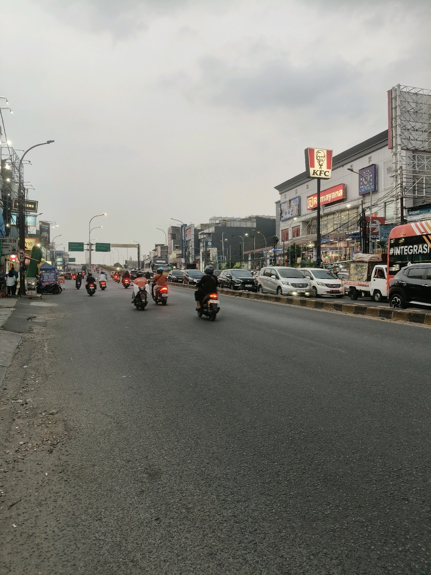 The image depicts a street scene in South Tangerang, Indonesia, on a cloudy day. A multi-lane road is filled with various vehicles, predominantly motorcycles, and some cars and trucks. Several individuals are riding motorcycles, some with passengers. On the right side of the road, buildings are visible, including a KFC restaurant with its iconic signage. Other businesses and billboards line the street. The road surface is asphalt, with a dirt and debris-filled shoulder on the left. Traffic appears to be moderate. The perspective is from street level, looking forward along the road, with the sky occupying the upper portion of the frame.