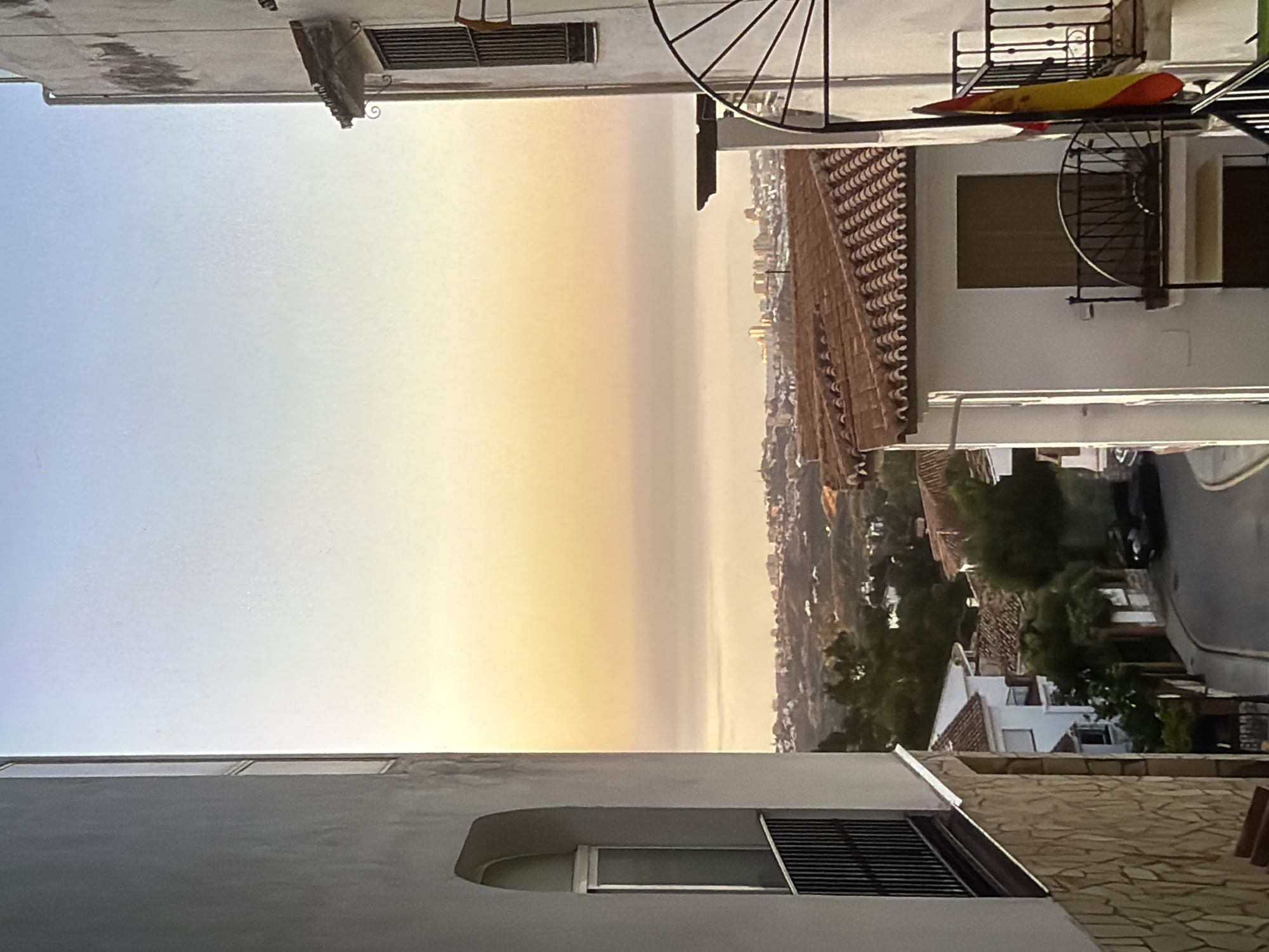 The media file captures a view from a high vantage point overlooking a coastal town at sunset. The sky is a gradient of soft blues and warm oranges, indicating the time of day. In the foreground, parts of buildings are visible, including concrete walls and tiled rooftops characteristic of Mediterranean architecture. A window with a metal grate is prominent on the left side.
To the right, a wrought-iron balcony features a Spanish flag, suggesting the location could be Spain. A spiral staircase is also visible on the balcony. Below the balcony, terracotta roof tiles are laid out in a traditional pattern. The landscape stretches out to the sea, which appears calm and blends with the hazy sky. A town with numerous buildings is spread across the hillside leading down to the coast. A winding street with a dark-colored car parked on it is also visible. The overall atmosphere suggests a peaceful, late evening setting.
