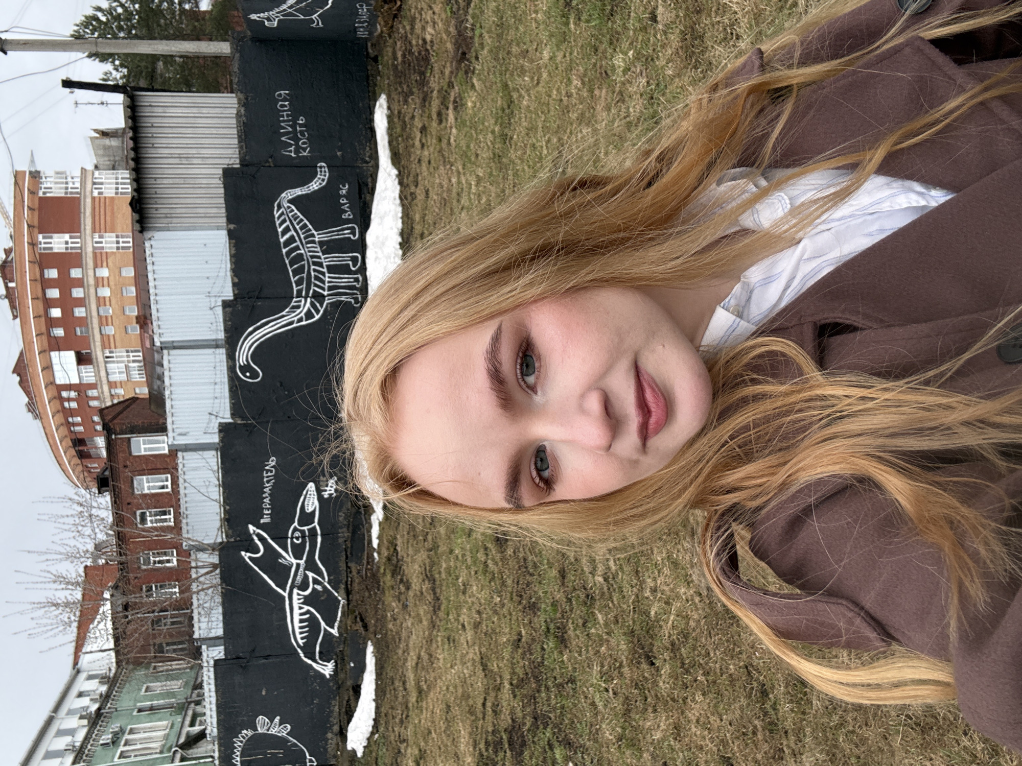 A photograph, taken in Perm, Russia, shows a fair-skinned, blonde-haired woman wearing a brown coat over a white shirt. She is positioned in front of a low, black brick wall adorned with white drawings of dinosaurs and Russian text. Beyond the wall is a grassy area and a view of buildings, including a multi-story brick structure. The sky appears overcast.