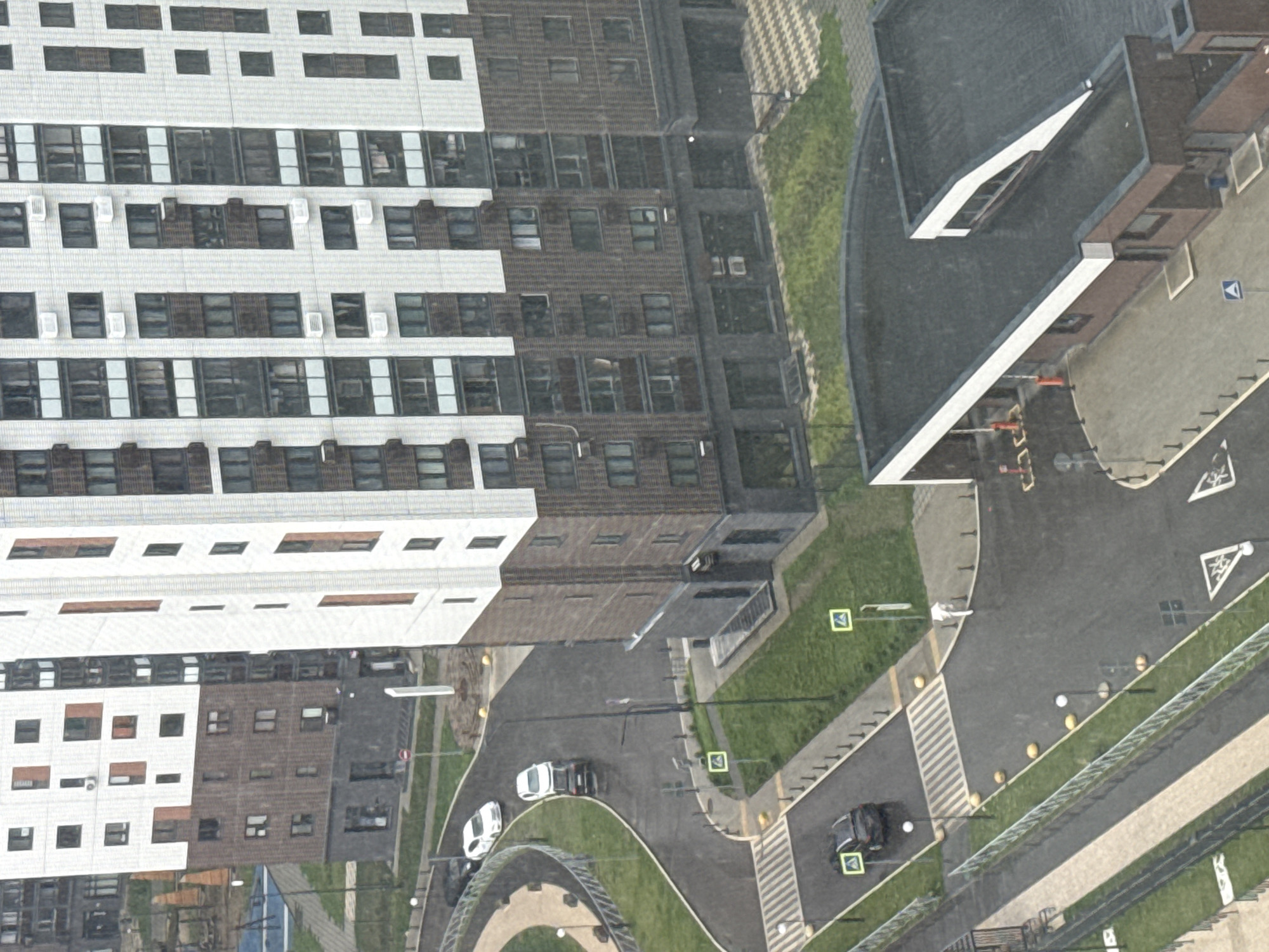 An aerial view shows a modern urban residential complex in Lyubertsy, Russia, on a rainy day. Several multi-story buildings with white and brown facades and numerous windows dominate the scene. One large building in the foreground features a pattern of alternating white and brown sections, with air conditioning units visible on the exterior. A smaller, darker building with a flat roof, possibly a commercial or service area, is situated in the lower right corner.
Roads and walkways wind through the complex. A black car is parked near a pedestrian crossing with white stripes, and a white car is driving on a wet road. Another black car is also visible on the road. Yellow bollards line the sides of the roads and walkways. Green grass patches are interspersed between the buildings and the paved areas. Several traffic signs, including a circular sign with a red border and a white horizontal bar, and square signs with green borders and white pictograms, are present. The rain is evident by the wet surfaces and the visible droplets in the air.
