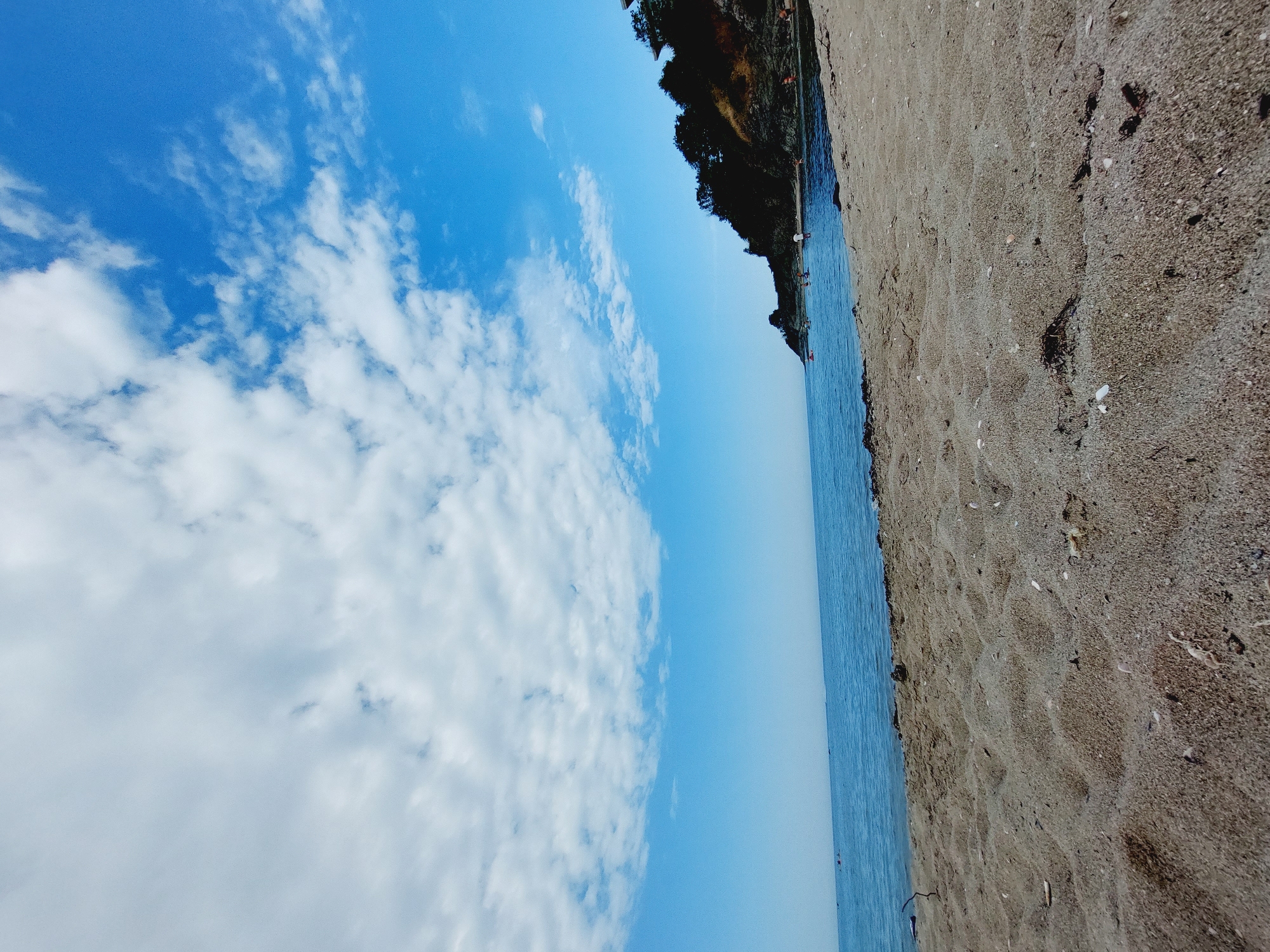 The image depicts a sandy beach with the Black Sea stretching out to the horizon under a partly cloudy blue sky. The foreground is dominated by textured sand with scattered shells and debris. The middle ground shows the calm, blue water with a few small boats in the distance. On the right side, a vegetated cliff face rises from the water, with some indistinct structures and people visible on it. Several individuals are also seen in the shallow water, appearing to swim or wade. The scene suggests a pleasant summer day at a coastal location, likely Kiten, Bulgaria, given the characteristic geography of the region.