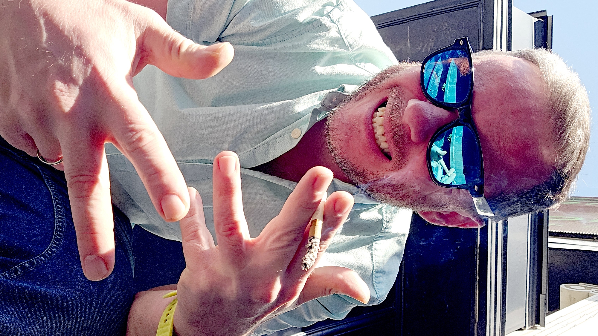 A person with light skin, fair hair, and a trimmed beard is facing the camera at a tilted angle, wearing reflective sunglasses. The reflection in the sunglasses shows a person holding up their hands. The person is wearing a light blue collared shirt. In the person's right hand is a lit cigarette with ash. The person is smiling, showing their teeth. A silver ring is visible on the left hand. A dark blue object, possibly clothing, is visible behind the left hand. A yellow wristband is also visible on the left wrist. A dark object, possibly part of a structure, is located behind the person. The location is unknown.