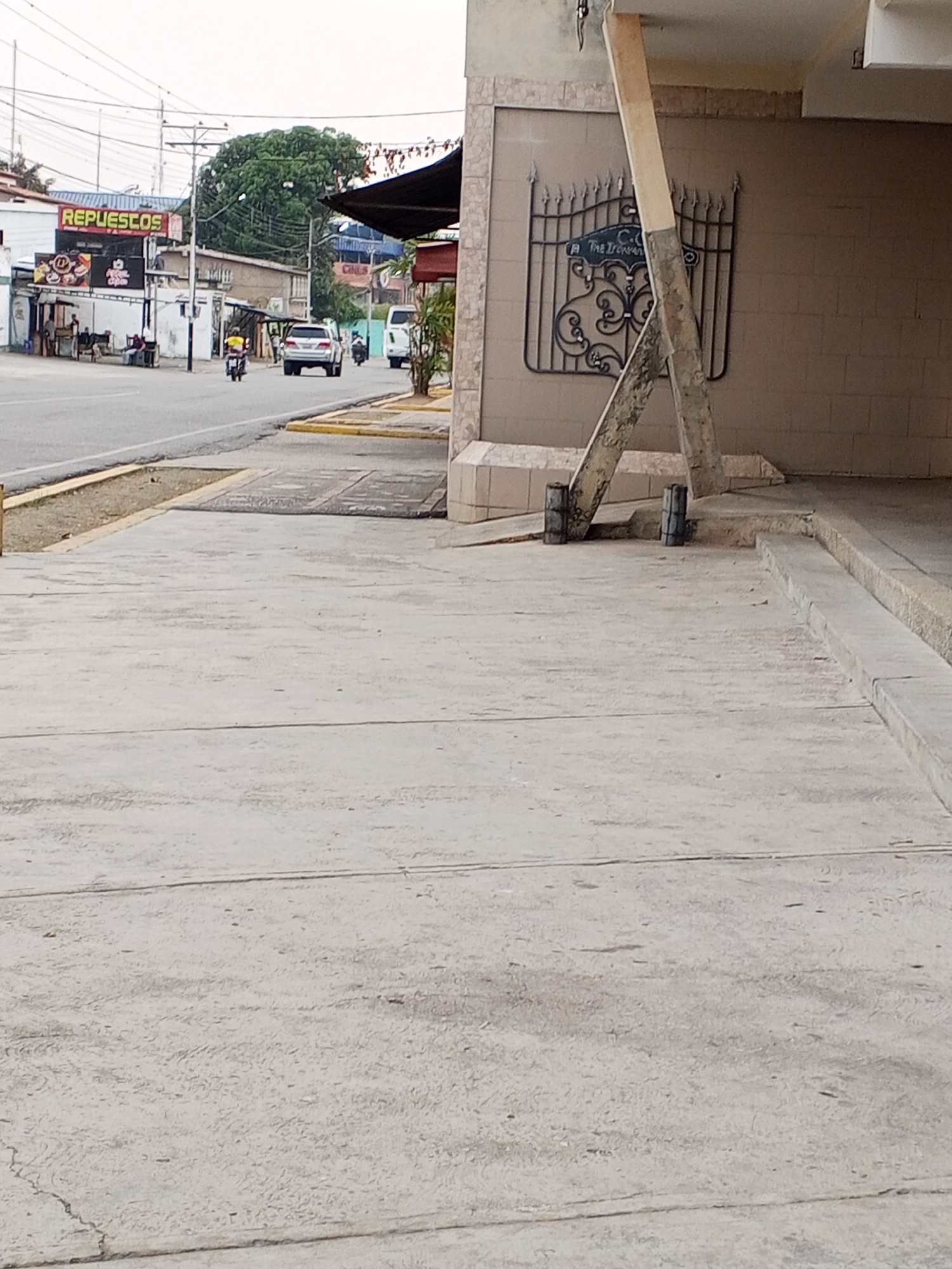 This is a daytime street scene captured in Tinaquillo, Venezuela.
In the immediate foreground on the right, a light-colored concrete sidewalk occupies the majority of the frame's lower half. To the right of the sidewalk, a building facade with tan-colored rectangular tiles is visible. A weathered, light-colored wooden beam extends diagonally from the upper right, across the tiled wall, to the ground, where two dark, cylindrical objects are positioned at its base. Affixed to the tiled wall behind the beam is a decorative black metal wrought-iron style piece, resembling a gate or grille, with a central blue sign bearing partially legible text "The Iron..." and "C.C.". Steps ascend from the sidewalk to an elevated section on the far right.
To the left of the sidewalk is an asphalt road. A yellow-painted curb separates the road from a narrow strip of dirt and sparse vegetation.
In the mid-ground, a silver SUV is observed traveling away from the viewer on the paved road. A person on a motorcycle, wearing a yellow shirt, is also moving away from the viewer, positioned to the left of the SUV. Further down the road, another motorcycle and a white utility van are partially visible.
On the left side of the street, a series of commercial buildings are present. The most prominent is a white structure featuring a large red sign displaying "REPUESTOS" (Spare Parts) in white lettering. Below this, a smaller sign with food-related graphics is attached. Approximately three to five individuals are gathered outside this building, near a seated arrangement.
The background reveals multiple utility poles heavily strung with electrical and communication wires. A large, dense green tree stands prominent behind the buildings on the left. The sky is overcast. The overall environment is urban and commercial.
