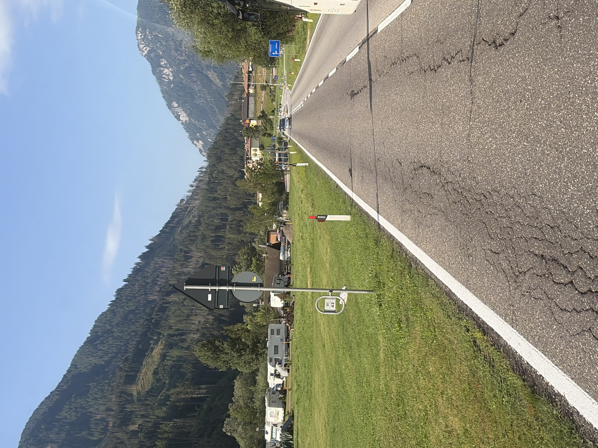 The image depicts a scenic road in Moena, Italy, flanked by lush green fields and towering mountains. On the left side of the road, a campsite is visible, with several recreational vehicles parked. A signpost stands in the field, indicating "VI 43". The road itself is paved and appears to be in moderate condition, with some cracks visible. Ahead, a few cars are driving on the road, which curves gently into the distance. The background is dominated by a majestic mountain range covered in dense evergreen trees. The sky is clear and blue, suggesting a pleasant day. The overall impression is one of a tranquil, picturesque setting in the Italian Alps.