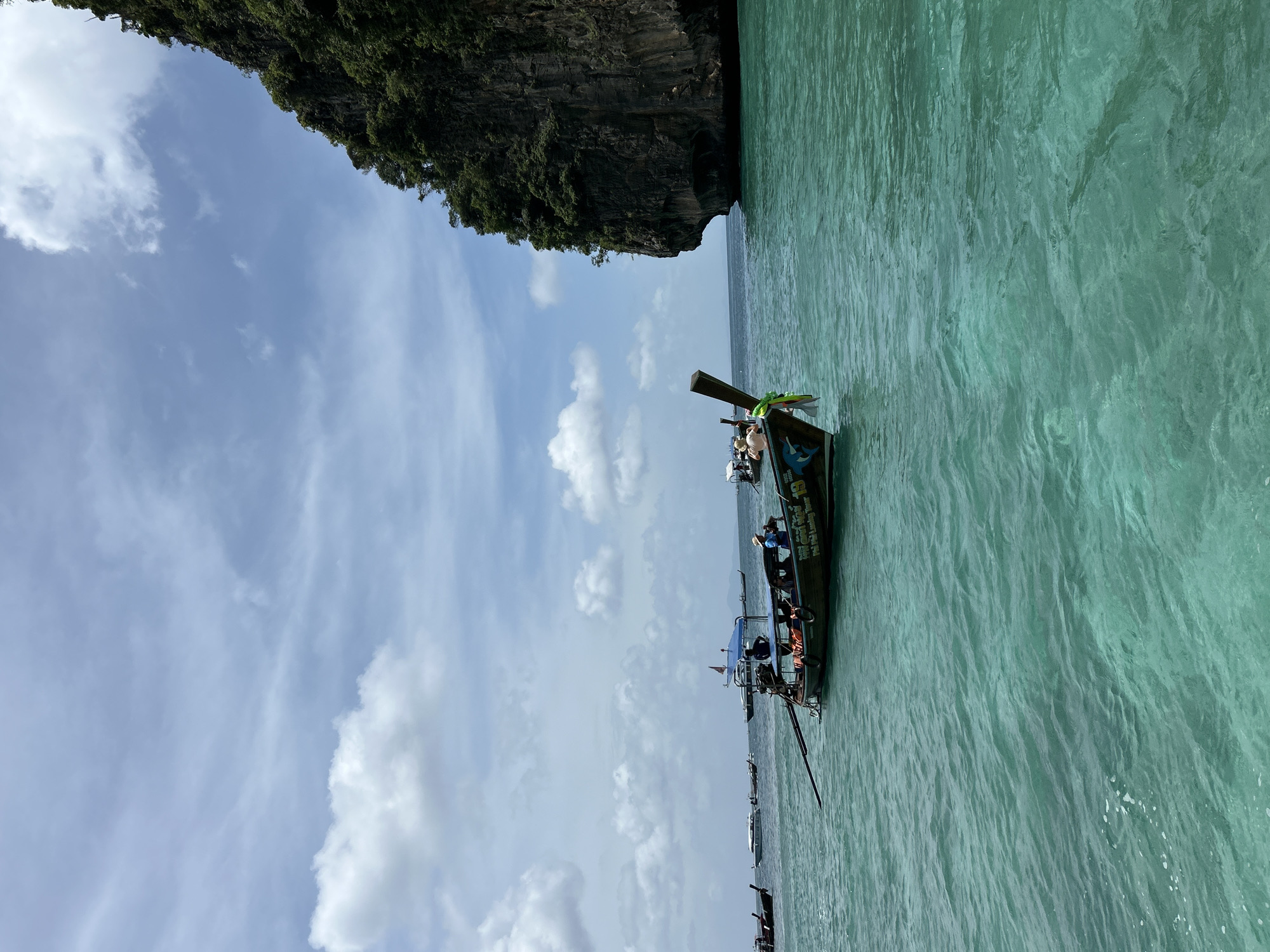 This image captures a serene view of the turquoise waters near Ao Nang, Thailand, on a partly cloudy day. In the foreground, a traditional longtail boat, characteristic of Thai waters, carries several passengers and is adorned with a decorative lei. The boat is positioned on the right side of the frame, with its wooden steering oar extending towards the left. In the background, a massive, lush green limestone karst rises vertically from the sea, a common geological feature in the region. Other longtail boats and speedboats are visible in the distance, suggesting this is a popular tourist area. The water is clear and calm, with gentle ripples indicating a light breeze. The overall impression is one of tropical beauty and tranquility.