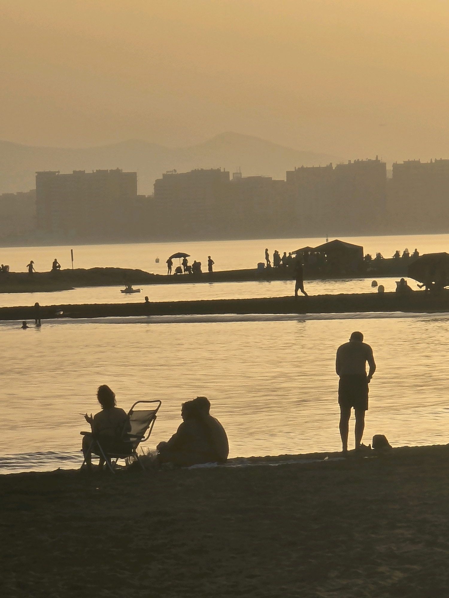 The image captures a serene beach scene in Málaga, Spain, during what appears to be sunset or a similarly hazy, golden hour. Silhouetted against the warm, diffused light are several groups of people.
In the foreground, a couple sits on the sand, one person in a beach chair and the other beside them. Further back, along a jetty or breakwater, a collection of figures are visible, some standing, others gathered around what might be a small structure or beach umbrella. A lone person walks along the same jetty, and further out on the water, a small boat is being paddled. In the middle ground, several individuals are in the water, with some submerged.
Across the expansive, calm body of water, a city skyline is discernible, characterized by numerous buildings forming a hazy silhouette against the horizon. The overall atmosphere is tranquil and relaxed, with the warm, muted tones of the light dominating the scene. The low angle of the sun creates long shadows and a soft, dreamlike quality.