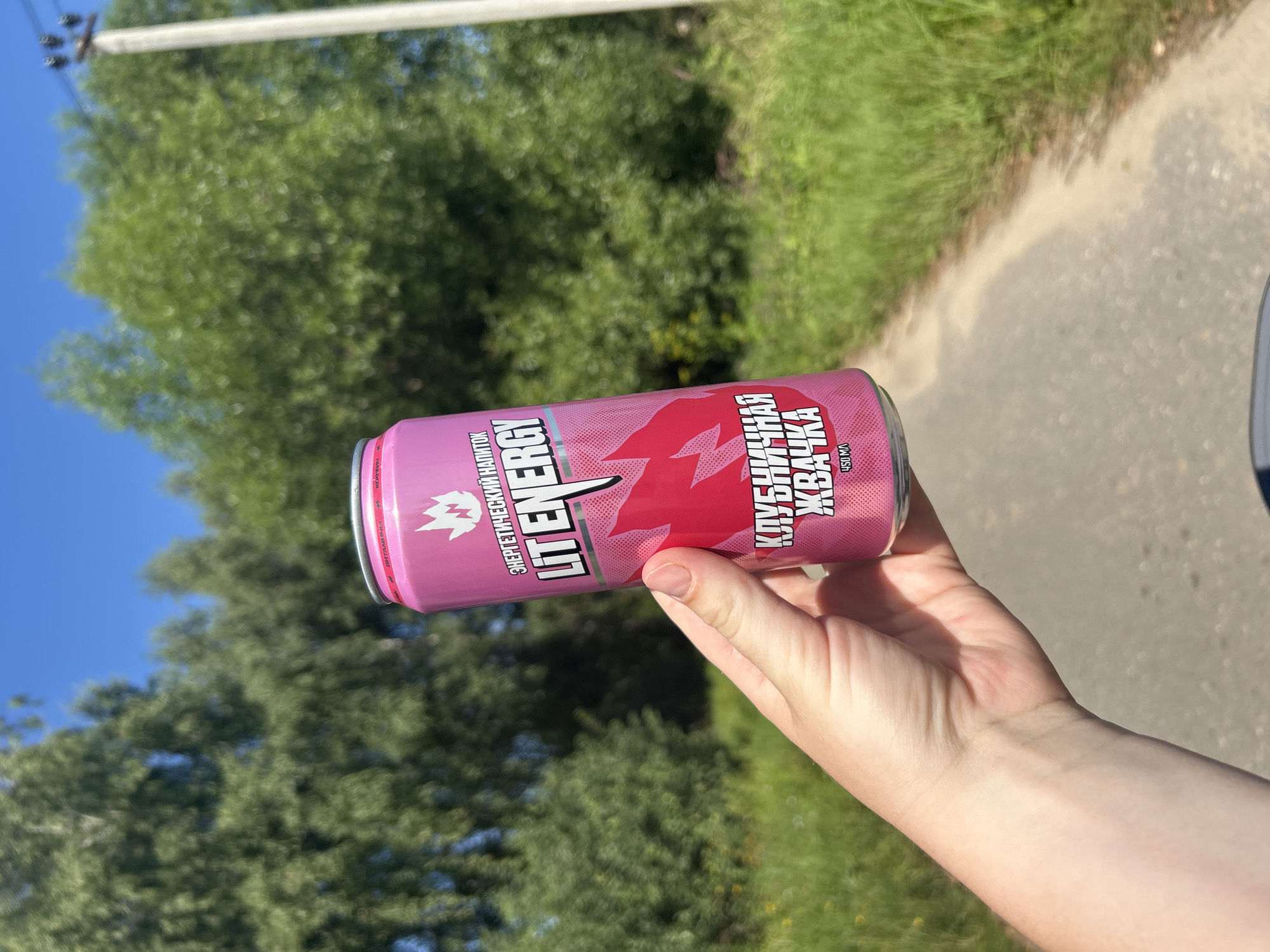A person's hand holds a pink can of "LIT ENERGY" drink with the flavor "КЛУБНИЧНАЯ ЖВАЧКА" (Strawberry Gum) against a blurred background of green trees and a blue sky. The can has a fire-like graphic and text in white and pink. The setting appears to be outdoors, possibly along a road, with tall grass and trees suggesting a rural or suburban environment. No specific landmarks of Kalitsino, Russia are visible.