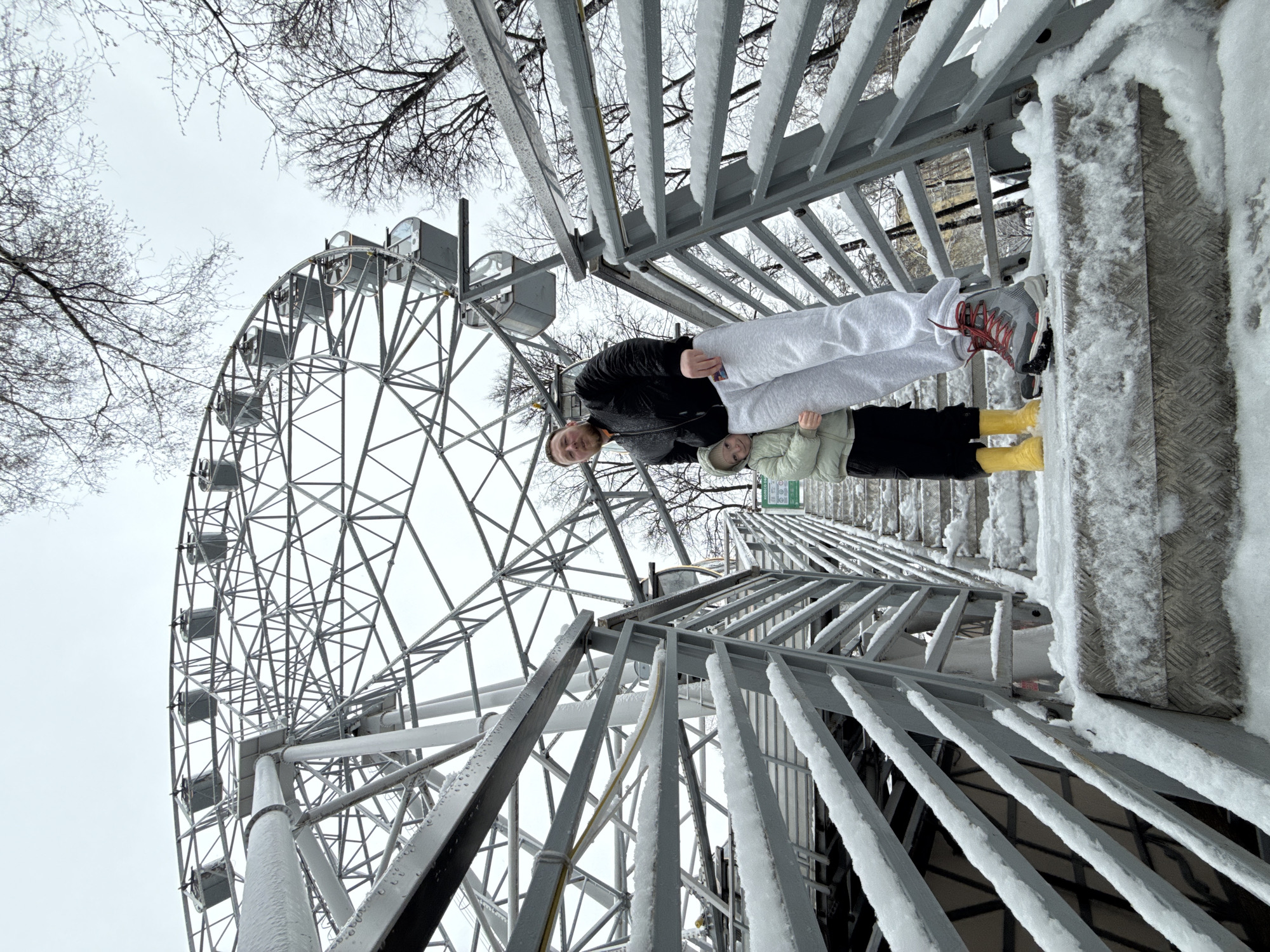 The image shows a man and a child standing on snow-covered metal stairs, near a Ferris wheel, on an overcast day. The Ferris wheel is a prominent feature, with its metal structure visible against the cloudy sky and bare branches of trees. The man is wearing a black jacket and light gray pants, while the child is dressed in a light green jacket and black pants, wearing yellow boots. The location appears to be in Perm, Russia, based on the style of Ferris wheel. The metal stairs are heavily coated in snow, and the overall scene suggests a cold, winter environment.