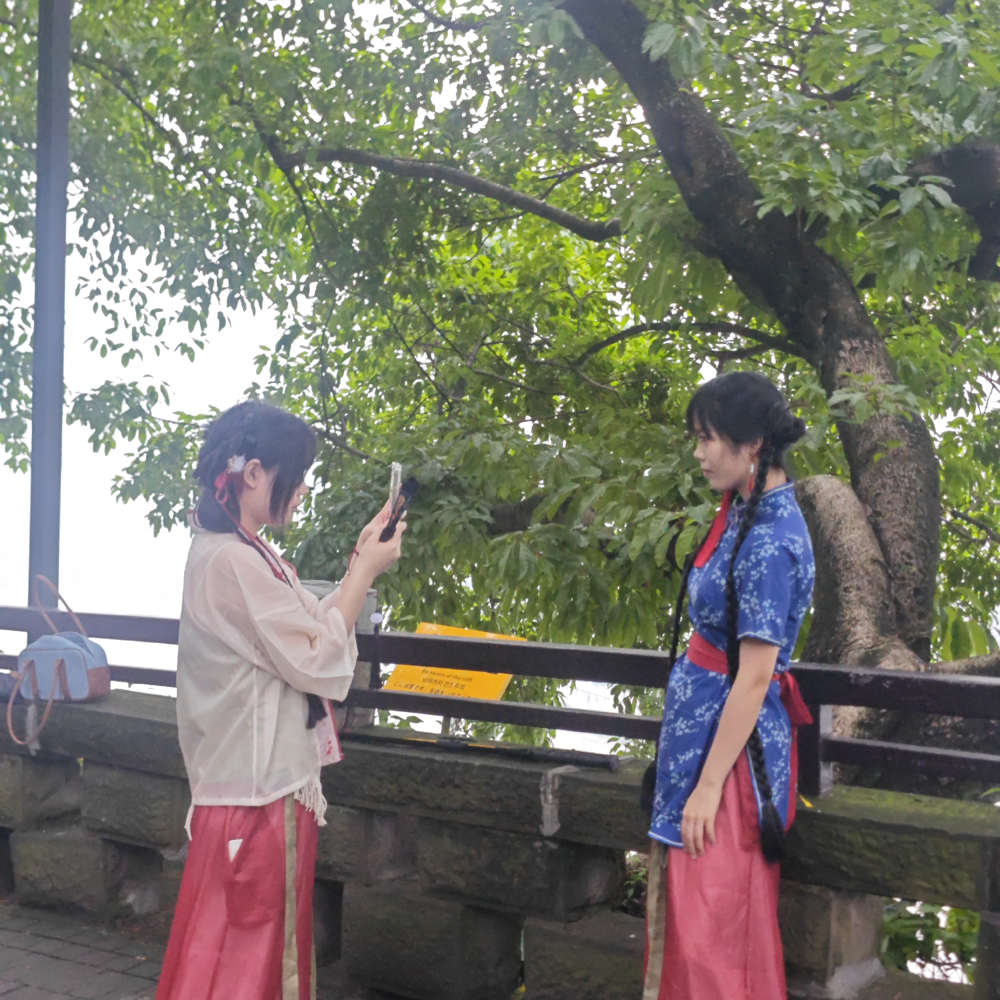 Two young women wearing traditional Hanfu clothing stand near a stone ledge. The woman on the left holds a mobile phone up, apparently taking a photograph of the woman on the right, who faces her with a gentle smile. A tree with lush green leaves rises behind them, partially obscuring the sky. A sign written in multiple languages warning of a cliff edge is visible on the railing between the women. A blue handbag rests on the ledge. The location is Yuzhong District, China.
