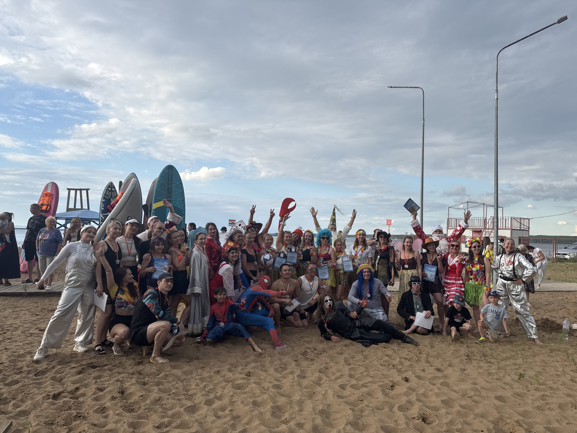 The image depicts a group of approximately 40-50 people, of varying ages and genders, posing for a photograph on a sandy beach. Many individuals are dressed in costumes, suggesting a festive or themed event. Some costumes include superhero attire (Spider-Man), traditional Russian folk costumes, a Santa Claus outfit, a spacesuit, and Hawaiian-themed outfits with leis and grass skirts. Several people are holding certificates or medals, indicating some form of competition or award ceremony.
In the background, a body of water is visible, likely a river or lake, with several paddleboards and other watercraft lined up on the shore. There are also two tall streetlights present, one on the left and one on the right side of the frame, which are typical urban street furniture. A low-rise building structure, possibly a pier or a recreational facility, can be seen in the background, situated near the water's edge. The sky is partly cloudy, with patches of blue sky visible. The overall setting appears to be a public beach area.
The location is identified as the Chaikovsky Beach in Chaikovsky, Russia. This beach is a known public recreation area situated on the Kama River, which is consistent with the presence of watercraft and the general landscape. The event appears to be a community gathering or a recreational activity involving water sports and costume participation.