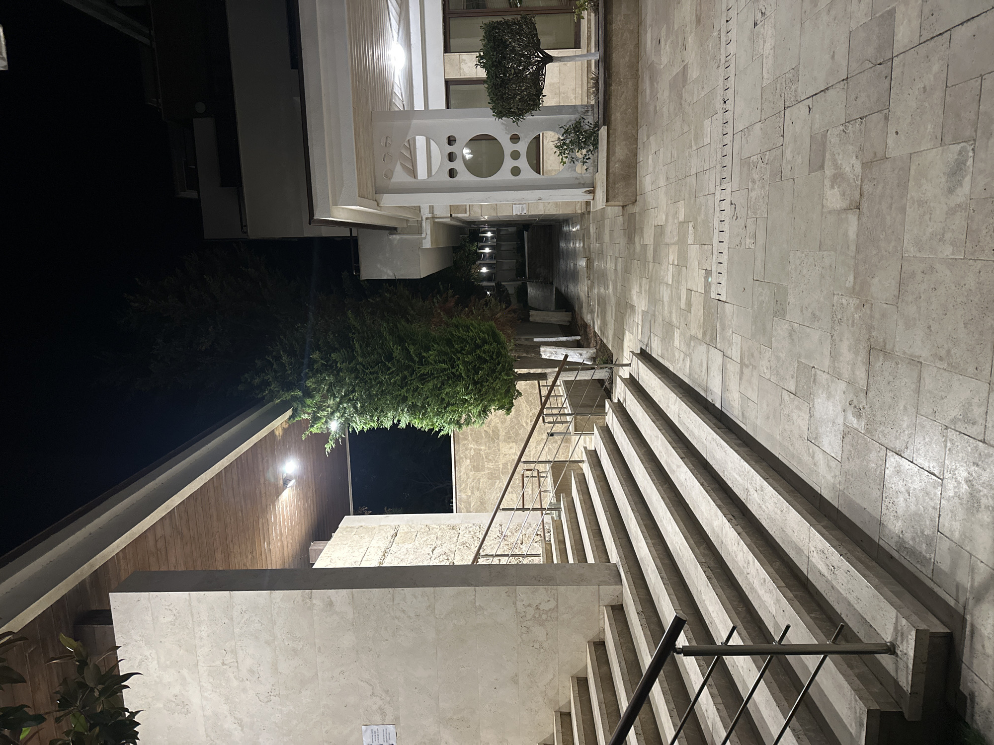 A nighttime exterior view showcases a paved walkway leading past a modern building with stone accents. On the left, a wide, multi-tiered staircase, constructed from light-colored stone, ascends towards an overhang with a wooden ceiling. A metal railing with wooden handrails lines the stairs. To the right of the stairs, a tall, dense evergreen tree stands. Further along the walkway, the building continues with a covered entrance supported by a decorative column featuring circular cutouts. Another small, topiaried tree is situated to the right of the column. The ground is paved with large, rectangular stone tiles, and a linear drain is visible in the walkway. The scene is illuminated by artificial lighting, casting shadows and highlighting the architectural features. The location is identified as Sozopol, Bulgaria.