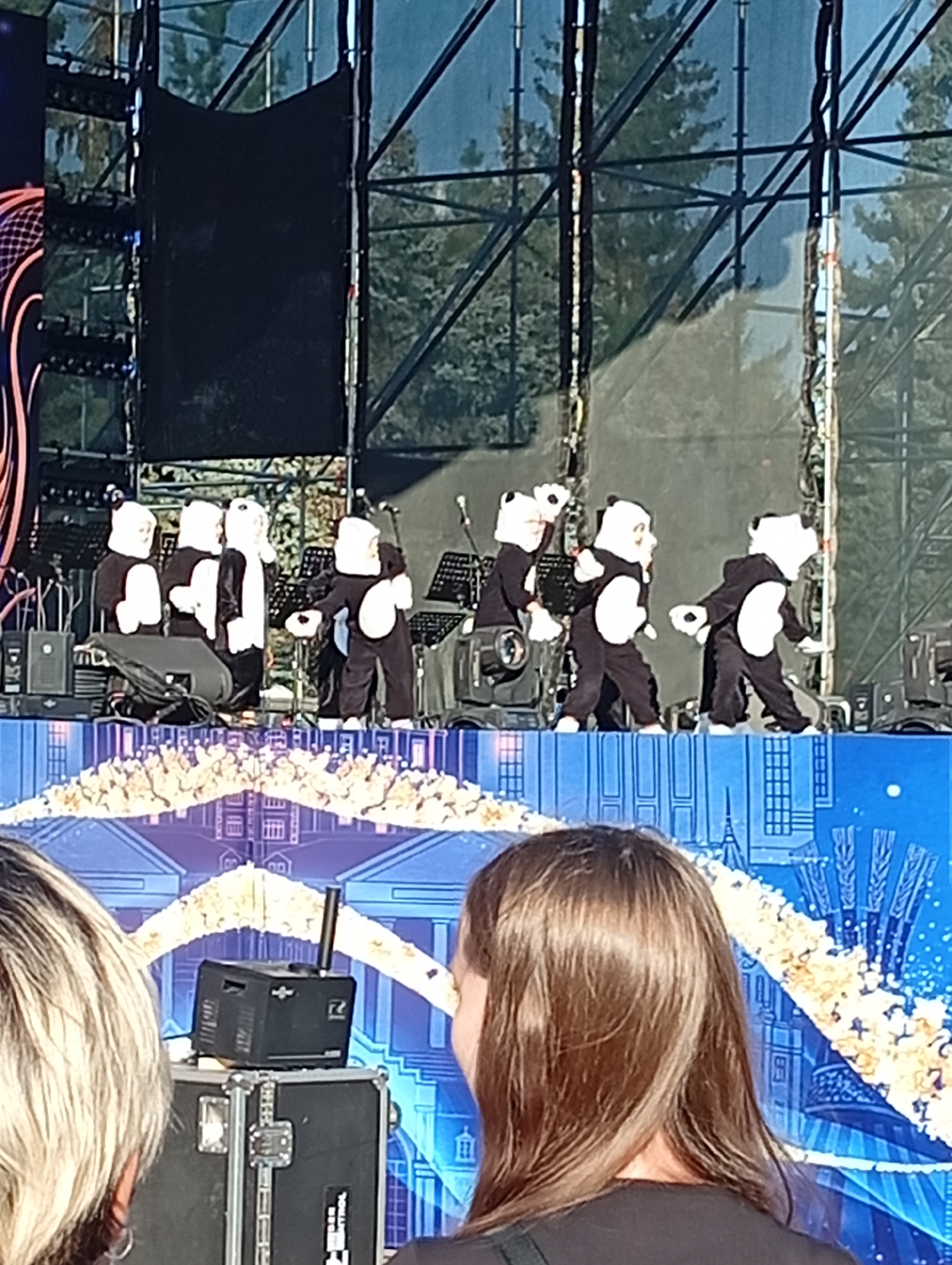 The image captures a performance on an outdoor stage in Kostanay, Kazakhstan. In the foreground, the backs of two spectators' heads are visible, one with blonde hair and the other with brown hair. The stage is adorned with a blue backdrop featuring architectural silhouettes, including what appears to be the Eiffel Tower, and a golden floral archway. The main focus of the image is a group of performers dressed in black and white panda costumes, standing on the stage. They appear to be engaged in a choreographed performance, with some of them interacting with each other. Several stage lighting fixtures, musical equipment including what looks like a speaker on a stand, and black fabric screens are also visible on the stage. The background reveals a metal truss structure and trees, suggesting an open-air venue.