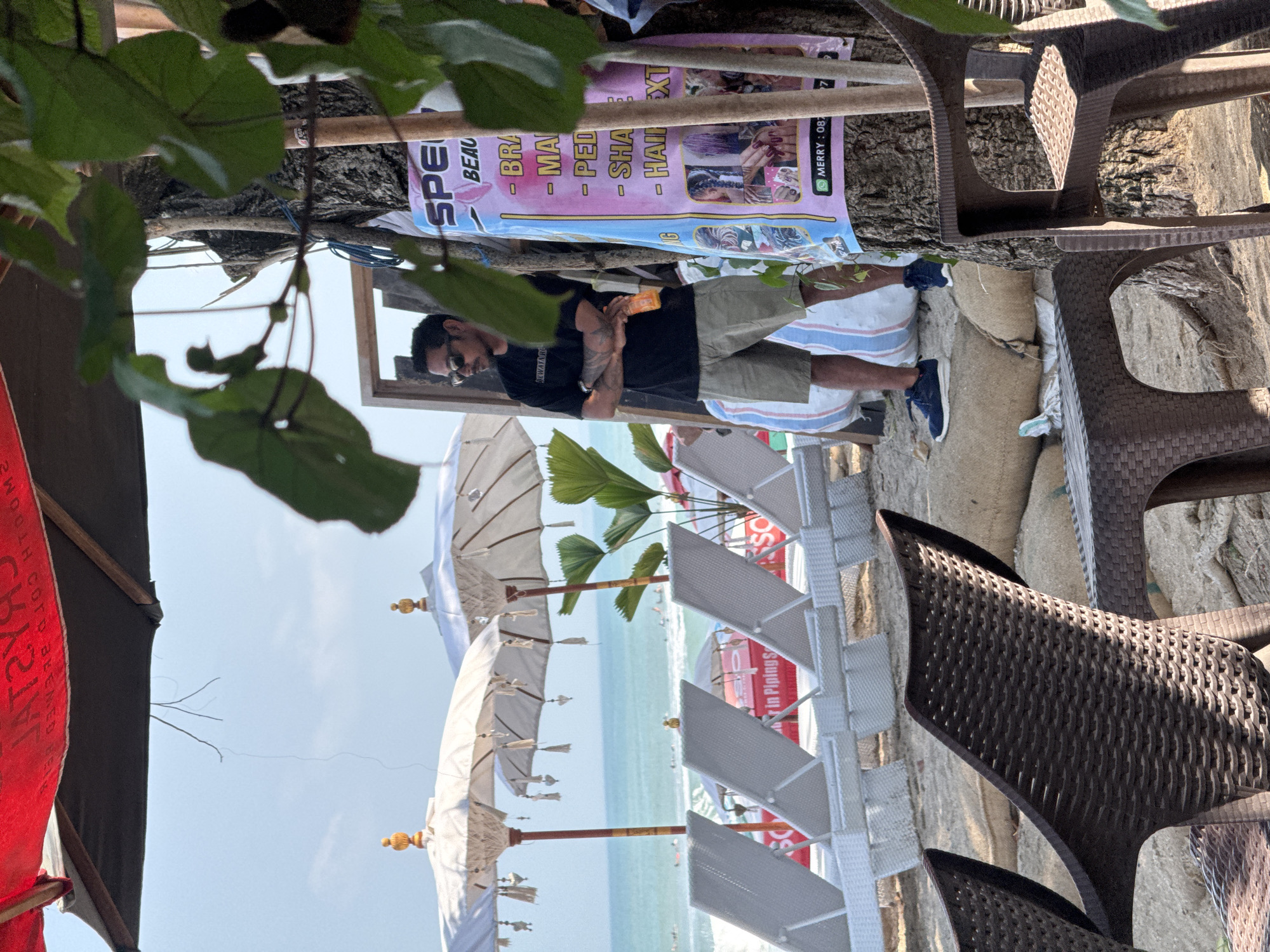 A man stands near a tree on a sandy beach in Kuta, Indonesia, with his arms crossed and holding an orange bottle. He is wearing a black t-shirt, khaki shorts, and blue sneakers. A poster advertising beauty services, including braids, manicures, pedicures, and hair extensions, is attached to the tree. In the background, beach chairs and umbrellas are set up near the ocean, with people swimming and surfing in the water. The overall scene depicts a relaxed beach atmosphere.