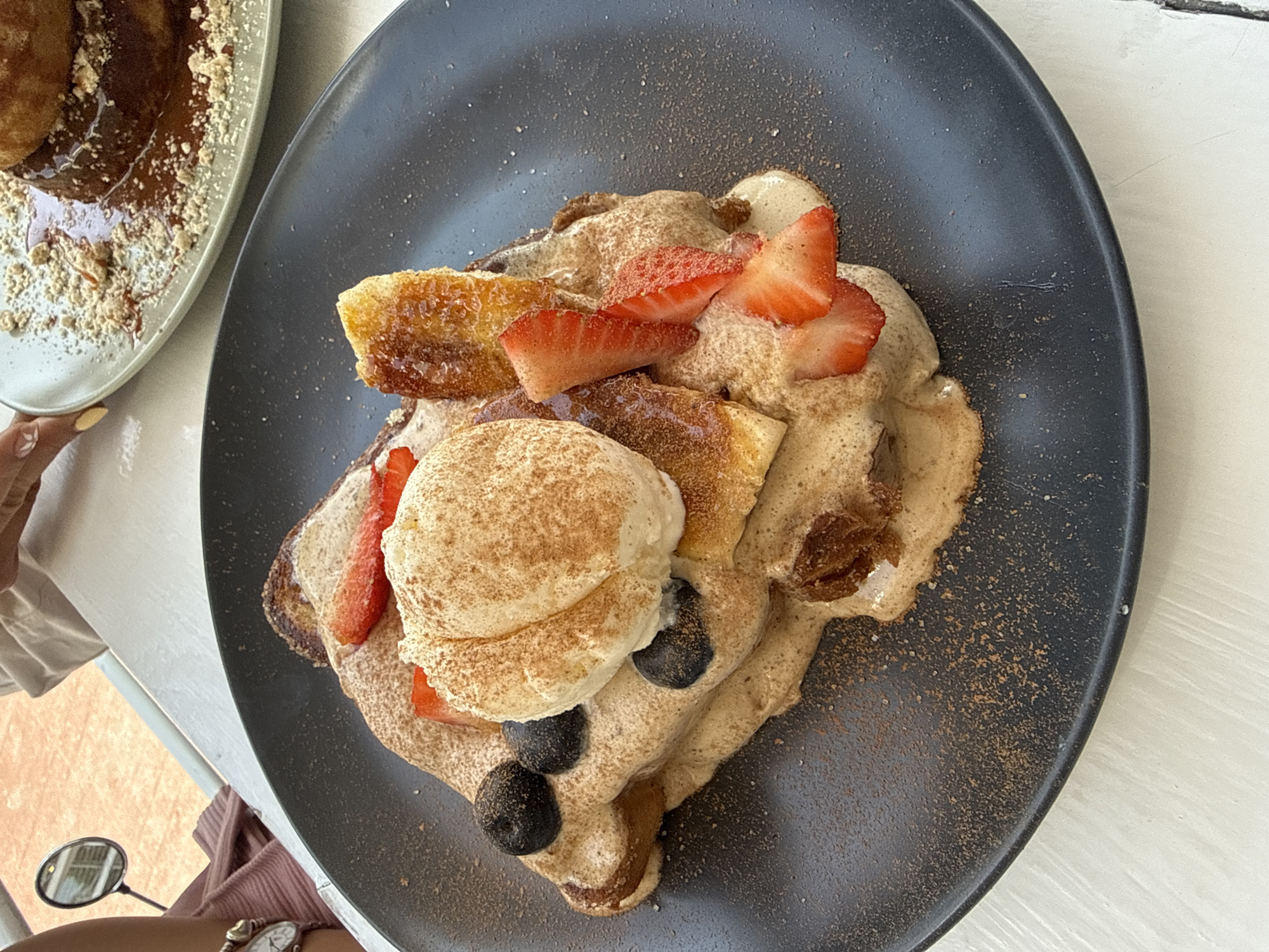 Here's a detailed description of the image:
**Scene:** The image shows a top-down view of a plate of food. It seems to be french toast with toppings.
**Food:** The main dish appears to be french toast, topped with what looks like a light brown sauce. There are slices of strawberries and grilled bananas. A scoop of white cream or ice cream is on top, sprinkled with a brown powder (cinnamon). There are also some blueberries.
**Setting:** The food is served on a dark gray, possibly matte, plate. To the left, a portion of another plate is visible, containing what appears to be dessert with a sauce and crushed nuts on top. The table is white and looks like it is indoors.
**Additional Objects:** A hand is slightly visible holding the plate from the left, the arm of the person is also visible with a watch. A purse, or similar accessory, is also visible.
**Environment:** Given the prompt about the location, it's presumed the photo was taken in San Andrés Cholula, Mexico. The environment implies an indoor setting, likely a restaurant or café.