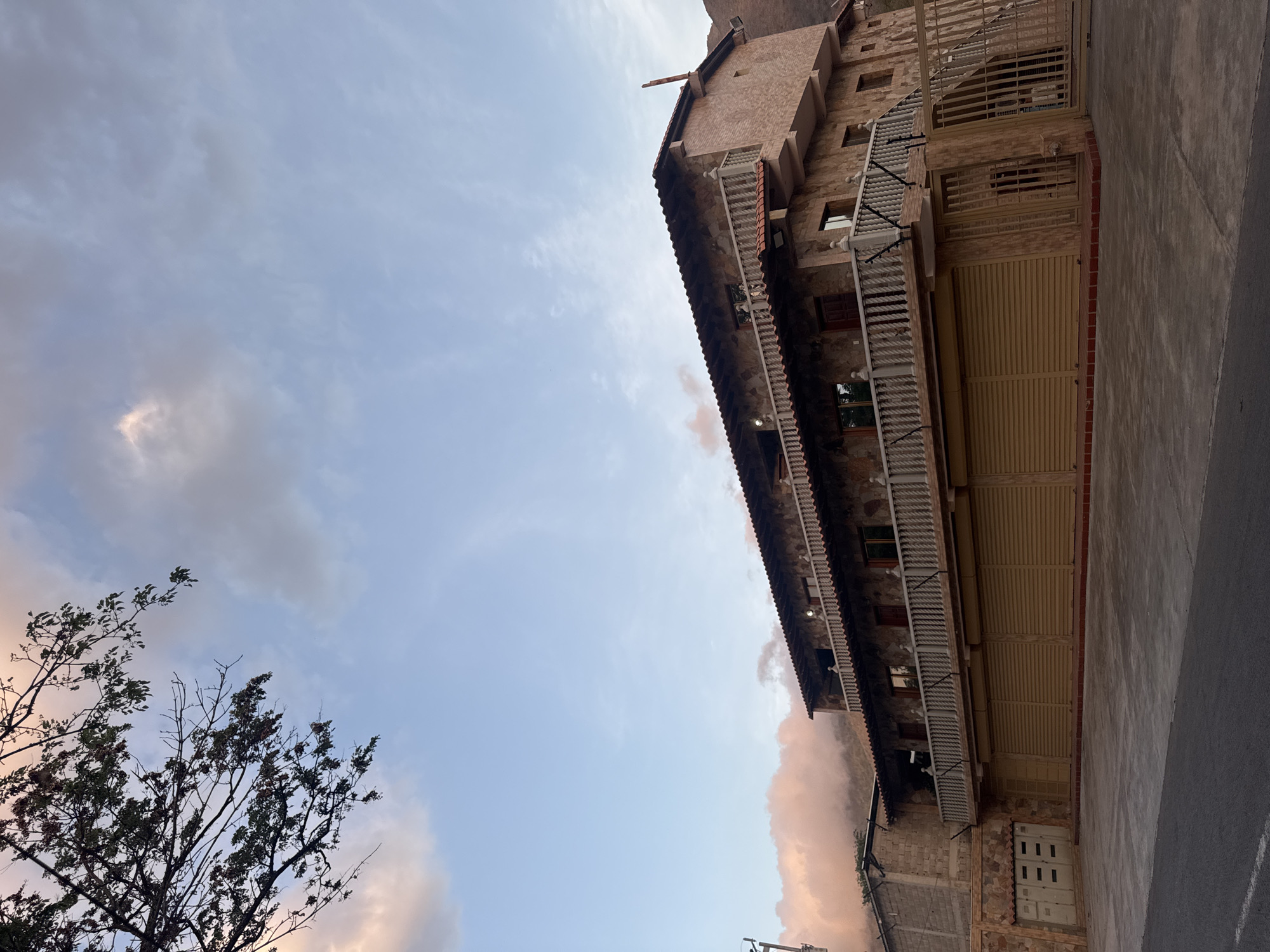 The image shows a multi-story building in San Juan Bautista, Venezuela. The building is constructed of stone and brick, with a red tile roof. There are multiple balconies on the front of the building, with white railings. The ground floor has a large, closed garage door. The sky is visible in the background, with some clouds. A bare tree is in the lower left corner of the image. A paved road is on the right side of the image.