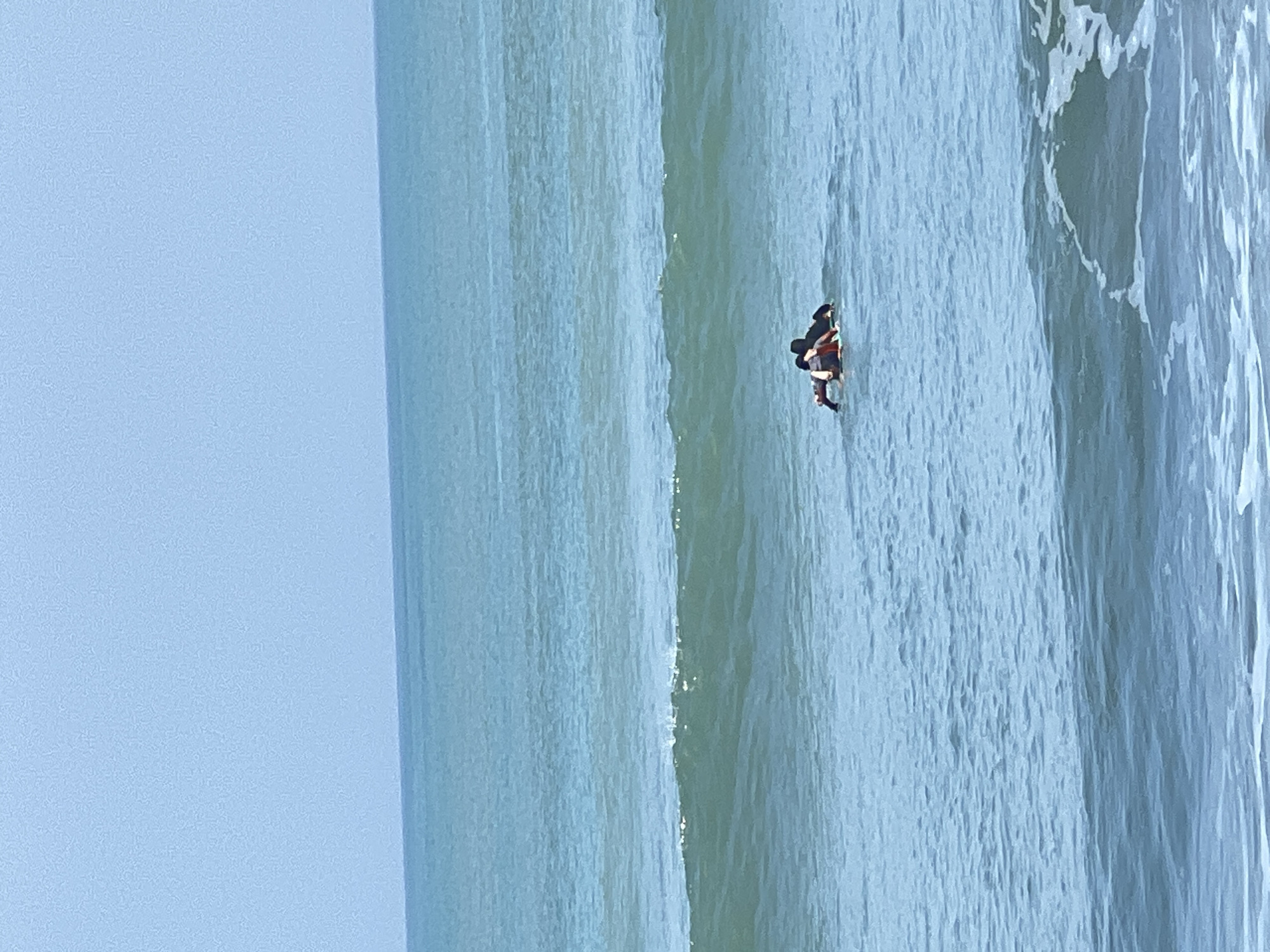 In the waters off Kuta, Indonesia, two individuals are engaged in surfing. One surfer, positioned further out, is lying prone on a surfboard, appearing to paddle or prepare for a wave. The second surfer, closer to the foreground, is also on a surfboard, facing away from the viewer and seemingly interacting with the first surfer, possibly assisting them. Both are in the ocean, with visible waves and the vast expanse of the sea stretching to the horizon under a clear sky.