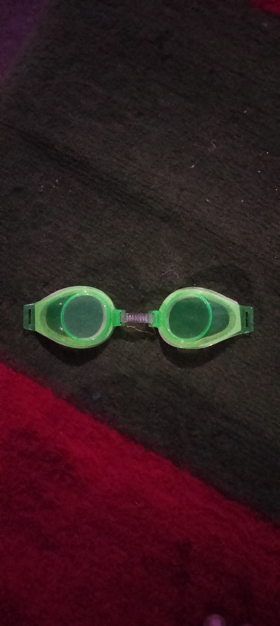In Serut, Indonesia, a pair of bright green swimming goggles lies on a dark, textured surface. The goggles are positioned horizontally in the center of the frame, with the lenses facing upwards. They are connected by a flexible, ribbed bridge. The background is a blend of dark, possibly fabric material, with a diagonal stripe of deep red visible at the bottom of the image. The lighting is dim, with some subtle reflections on the goggles.