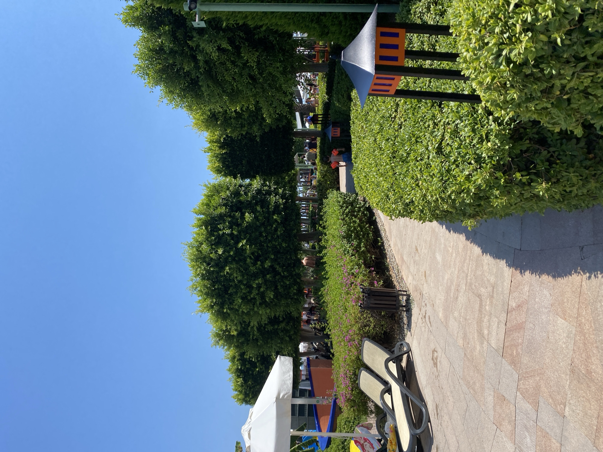 The image depicts a sunny day at what appears to be a resort or park area in Aksu, Turkey. In the foreground, a paved pathway made of rectangular stone tiles is visible, bordered by a neatly trimmed green hedge. Two empty lounge chairs are positioned on the left side of the pathway, under a white umbrella. Further into the scene, a row of lush green trees forms a natural canopy. Behind the trees, several people can be seen scattered throughout the area, some appearing to be children playing. In the middle ground, a low hedge with small pink flowers is present, and a dark metal trash bin stands beside the pathway. A decorative lantern with orange and blue panels sits on a black stand in the right foreground, partially obscured by the hedge. The sky is a clear, vibrant blue, indicating fair weather. The overall impression is that of a well-maintained outdoor recreational space.