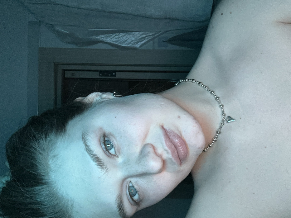 The image presents a close-up portrait of a female individual captured in an indoor setting. The precise location within Noviy, Russia, is not discernible from the visual evidence; however, the background elements suggest a private or residential interior, possibly a bedroom or living space.
The subject is a young adult female with light-colored skin, light blue eyes, and natural-looking eyebrows. Her dark hair appears pulled back or styled upwards. Her lips are full, and her skin exhibits visible pores and minor imperfections, consistent with natural texture. She is looking directly towards the viewer.
She appears bare-shouldered from the visible portion, wearing a silver-colored choker-style necklace composed of small, uniform beads. A small, pointed, heart-shaped pendant hangs centrally from the necklace. A silver-colored earring, possibly a small hoop or stud, is visible in her right ear (viewer's left).
The background is dimly lit and largely out of focus, exhibiting a cool, bluish-green ambient light. On the right side of the image, a vertical section of a door frame and visible dark hinges suggest an open door or a structural opening. Further right, an indistinct light-colored fabric item, potentially a curtain or hanging garment, is partially visible against a dark wall. No other individuals or explicit actions or interactions are depicted. The scene is illuminated by an artificial light source contributing to the predominant cyan color cast across the subject and the immediate environment.
