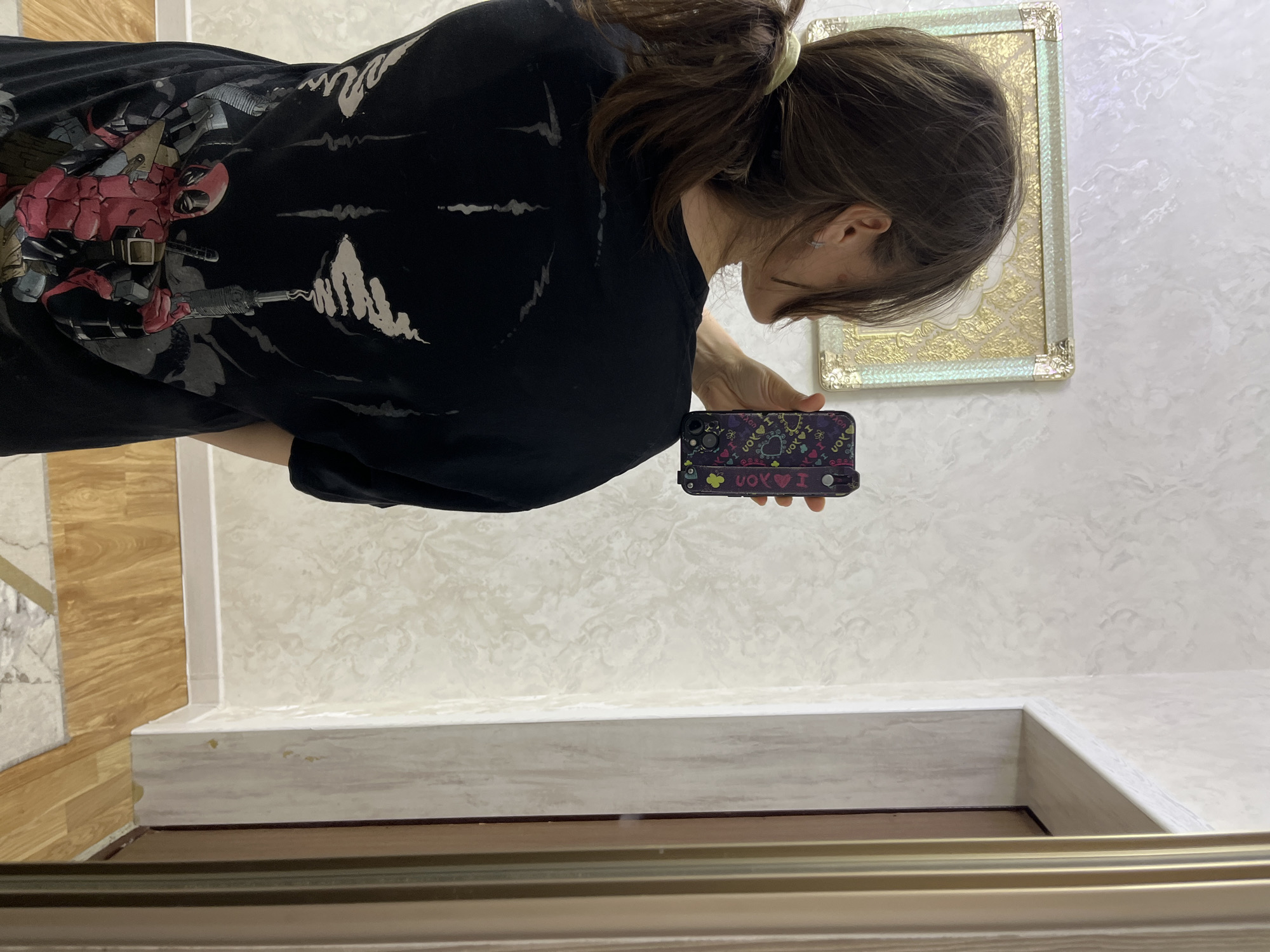 A young woman with dark brown hair tied in a ponytail is seen from behind, taking a mirror selfie. She is wearing a black t-shirt with a graphic print of Deadpool on the back. Her right hand is holding a smartphone, which is also facing the mirror. The background consists of a textured wallpaper, a framed picture on the wall, and a doorway. The floor is made of wooden planks and part of a rug is visible. The image appears to have been taken indoors, possibly in a residential setting. There is no identifiable information in the image to suggest the location is the city of Kaspiysk, Russia.