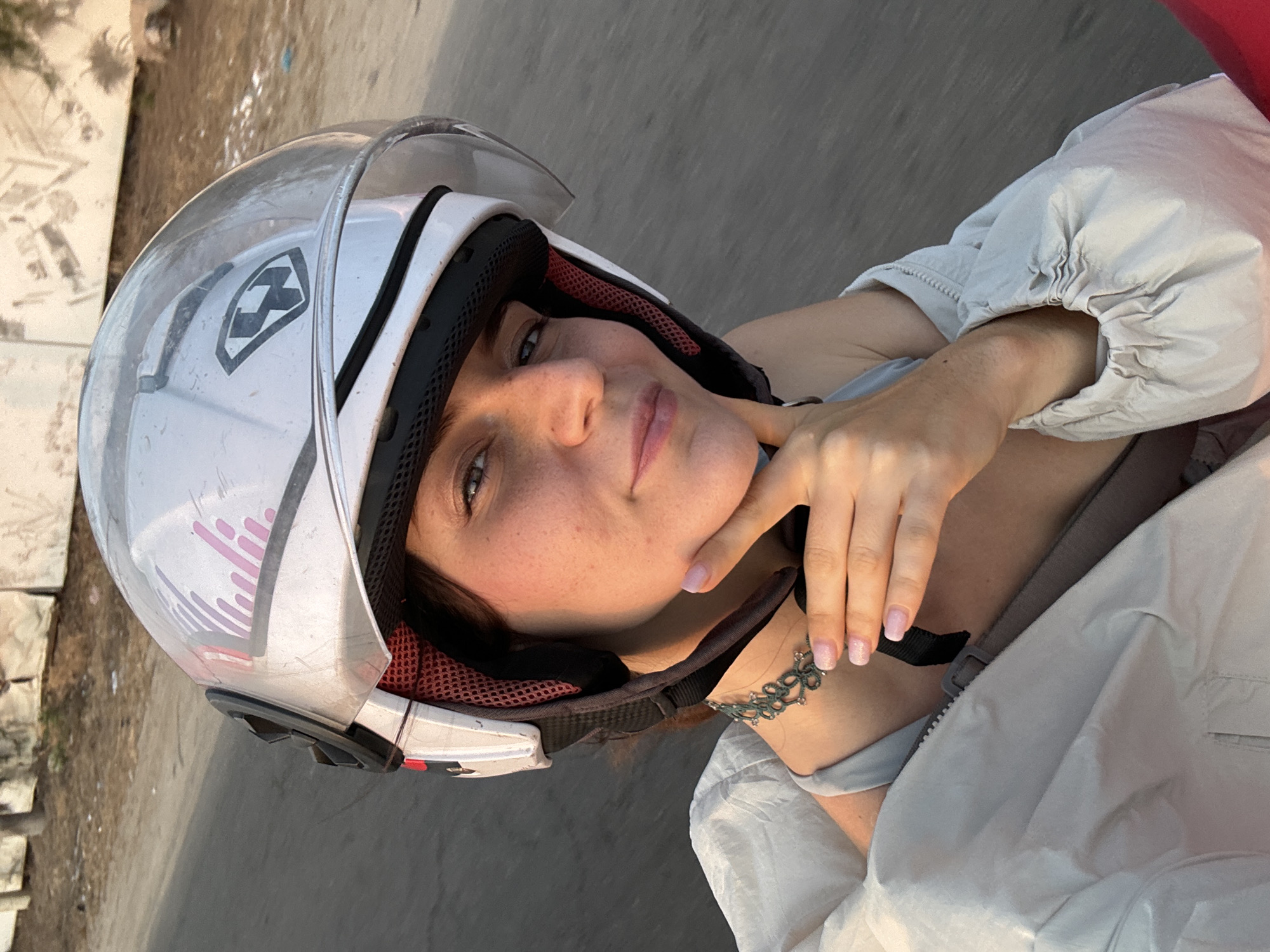 A woman wearing a white helmet with a clear visor and pink and gray detailing is the primary subject of this selfie-style photograph. She is wearing a light-colored jacket and has her hand near her chin. Visible is a black choker necklace. The background shows a street, and the photo is likely taken in the city of Mũi Né, Vietnam.