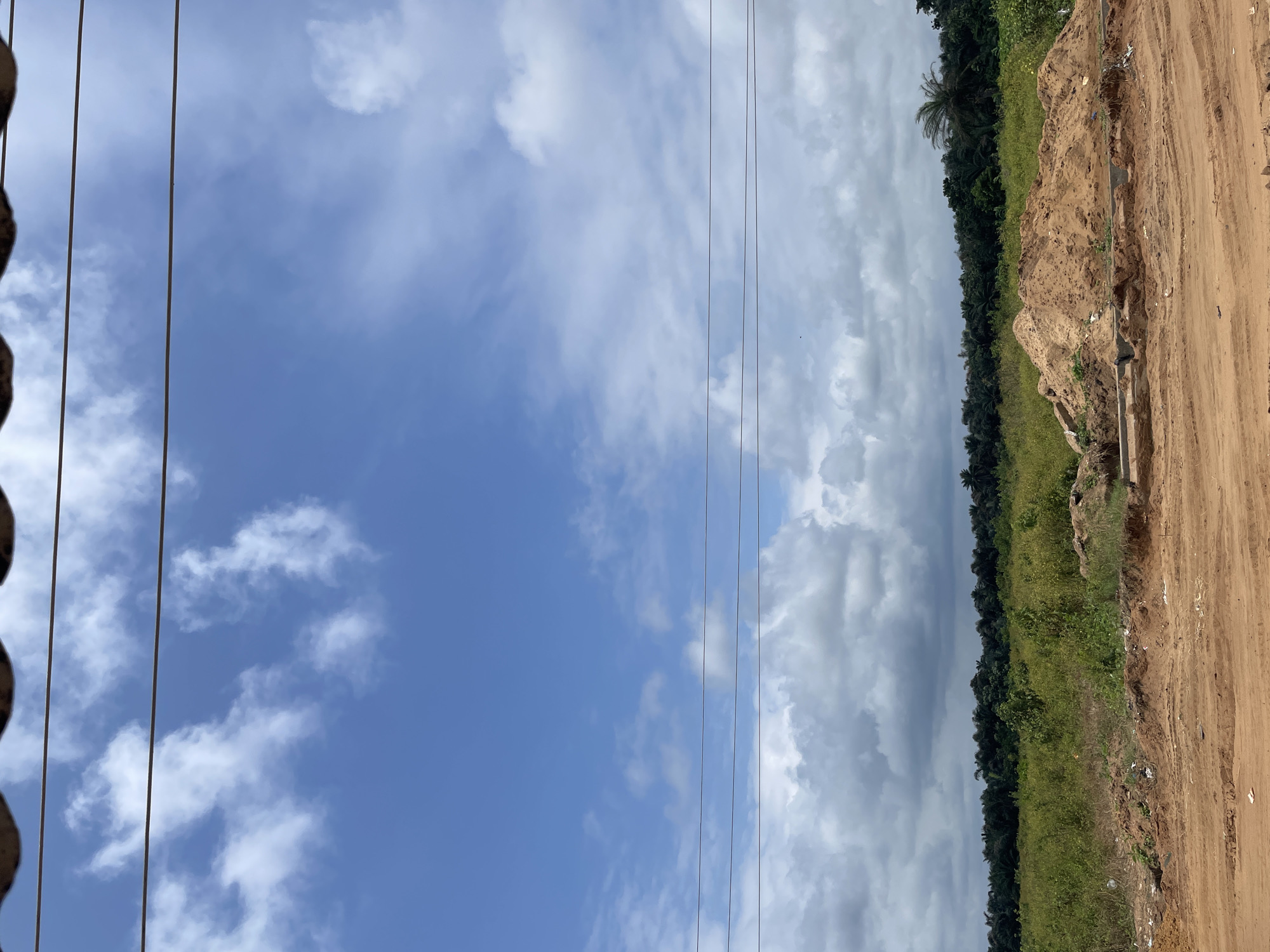This image depicts an outdoor scene in a rural area, likely in Iyafin, Nigeria. The foreground is a dirt road with tire tracks, leading towards a construction site where a large pile of sand sits to the right. Beyond the sandpile, a field of green vegetation stretches towards a dense line of trees in the background. The sky is a vibrant blue with scattered white clouds. Three thin, dark power lines are strung horizontally across the upper portion of the image, disappearing into the trees. The overall impression is one of an undeveloped or developing area with elements of both nature and human activity.