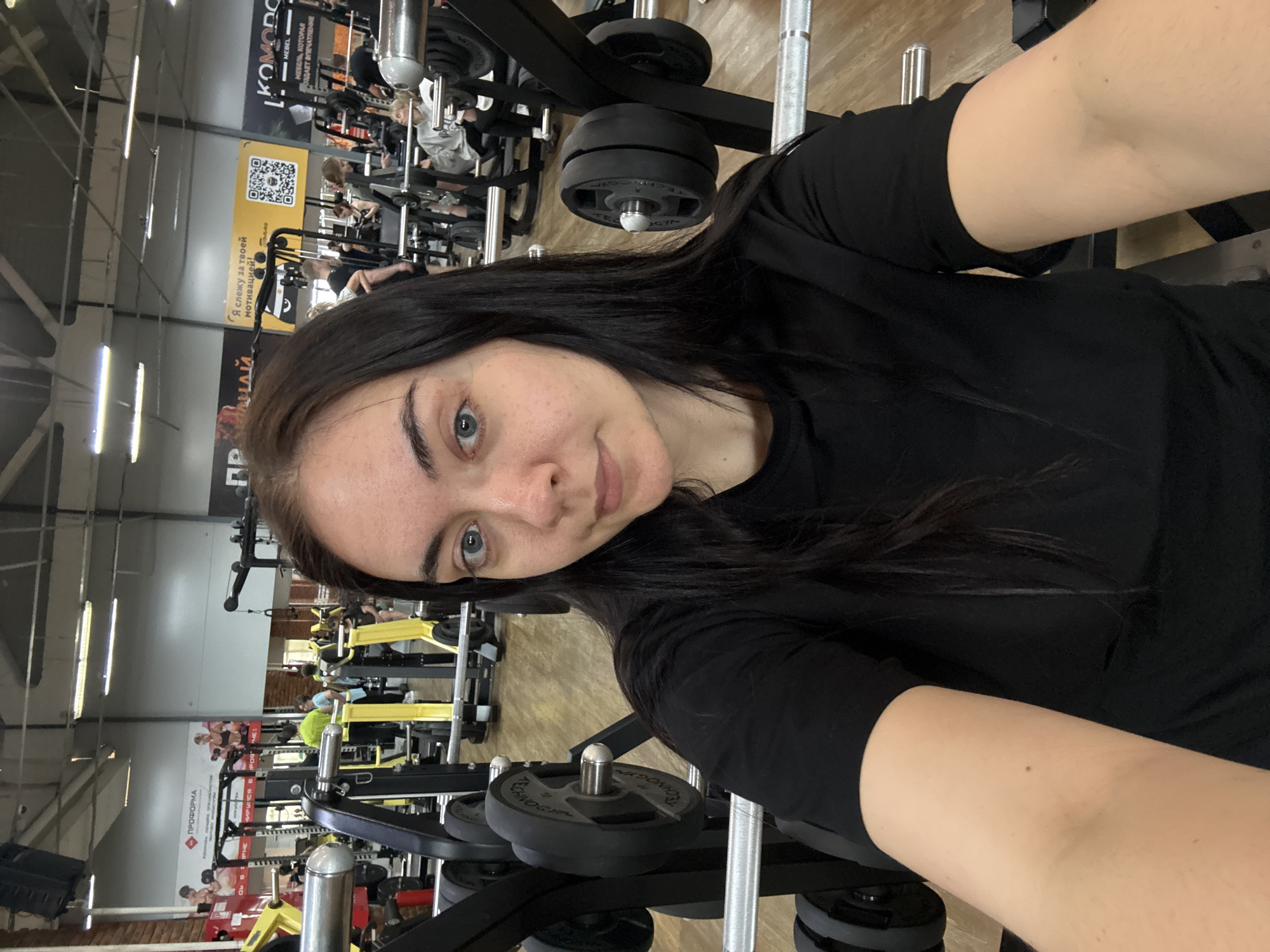 The image captures a woman in a gym setting, likely in Chaikovsky, Russia, based on the Russian text visible in the background. She has dark hair and light eyes, and she is wearing a black t-shirt. The gym is visible in the background, with various weightlifting machines and other equipment. Other individuals are present in the background, engaged in their own workouts. The text "KOMODO" and "ПРОФОРМА" can be seen on the gym walls. The picture seems to be a selfie taken with the woman holding the camera, as the angle suggests. The gym appears to be well-lit with overhead fluorescent lighting.