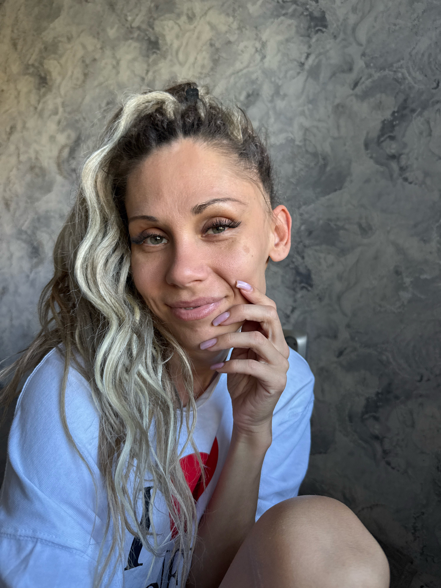 A woman with blonde and dark curly hair pulled back and a white t-shirt with a red heart graphic is shown sitting in front of a textured grey wall. Her right hand is raised to her cheek, with her fingers resting on her chin. Her fingernails are painted a light pink color. Her gaze is directed towards the camera with a slight smile. The lighting suggests an indoor environment, possibly in Zhukovo, Russia, with natural light coming from the left. The background appears to be a wall with a mottled, concrete-like texture.