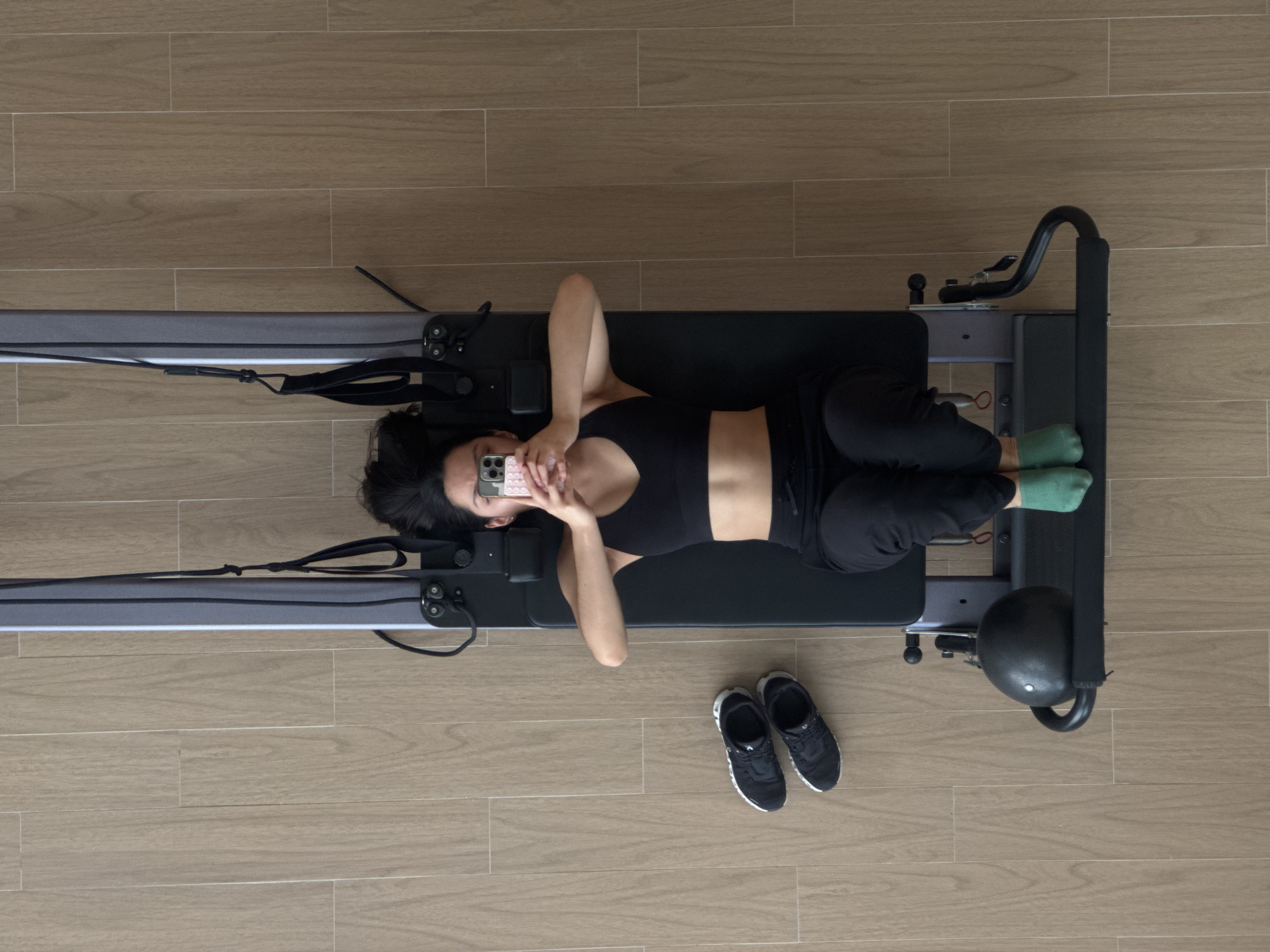 A top-down shot shows a woman lying on a Pilates reformer machine in a room with light brown, wood-look flooring. She is dressed in a black sports bra and black athletic pants, with green socks visible on her feet. She is holding a pink cell phone in front of her face. Next to the machine is a pair of black sneakers. The location is San Bernardino Tlaxcalancingo, Mexico.