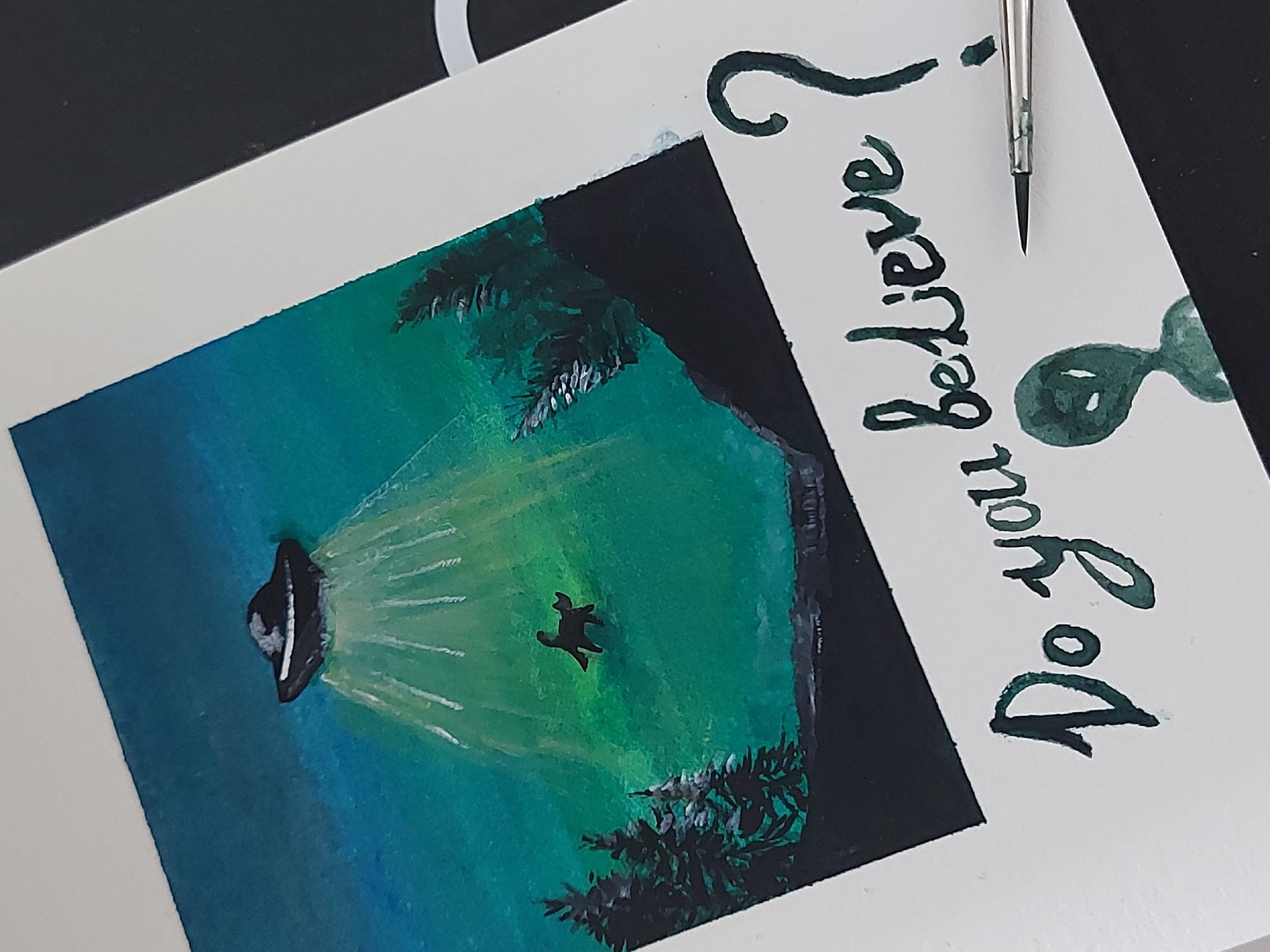 The image is a close-up of an artwork depicting a UFO abduction scene. The artwork shows a black UFO shining a beam of white light down upon a figure, presumably a human, floating in the beam. The environment is painted in shades of blue and green, with dark trees visible in the foreground and along the sides of the scene. The words "Do you believe?" are hand-painted in green ink to the right of the scene. A paintbrush is also visible, resting on the artwork near the question mark. The city of Chaikovsky, Russia is not apparent.