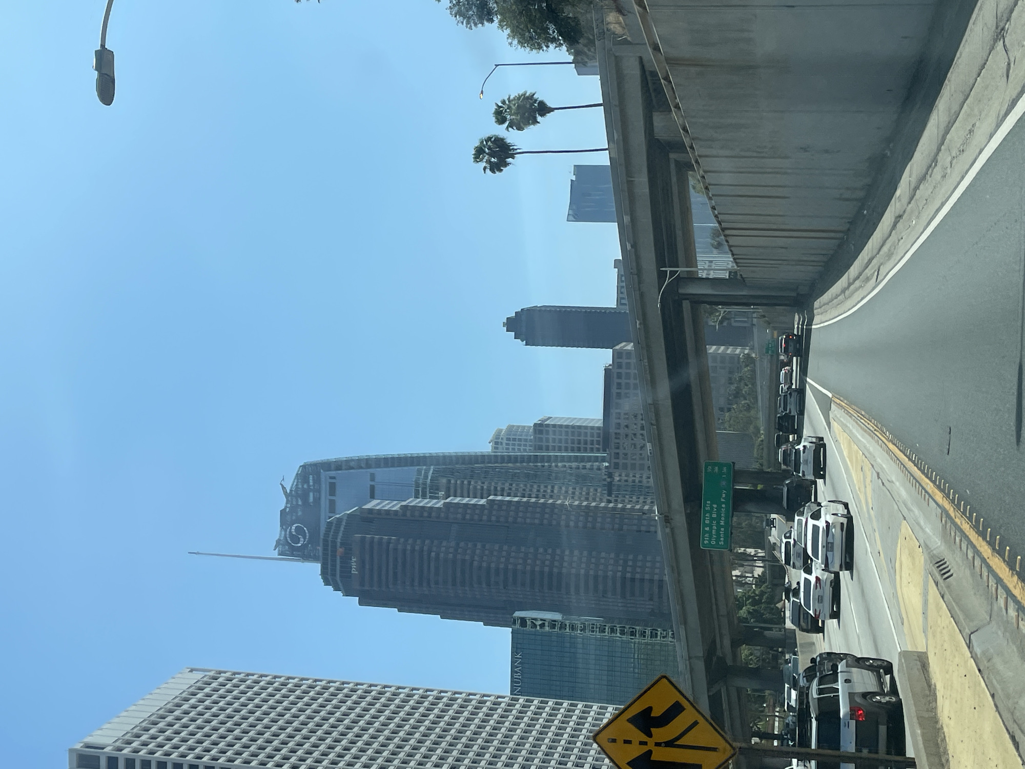 The image depicts a daytime scene from the perspective of a vehicle traveling on a multi-lane highway, looking towards a city skyline under a clear blue sky.
In the immediate foreground, to the left, a yellow diamond-shaped road sign with black arrows indicates merging lanes. A low concrete barrier runs along the left edge of the road, separating the shoulder from the traffic lanes. Several vehicles, including a white Honda Element and multiple sedans, are visible on the road to the left.
The midground features a multi-lane highway with moderate to heavy traffic, consisting of various cars and SUVs moving in the same direction. White and yellow lane markings are clearly visible on the asphalt. An elevated concrete freeway structure spans horizontally across the scene. Mounted below this structure is a green overhead road sign indicating exits for "9th & 8th Sts", "Olympic Blvd", and "Santa Monica Fwy".
In the background, beyond the highway infrastructure, the urban landscape of downtown Los Angeles is visible. Prominent skyscrapers include the Wilshire Grand Center, identifiable by its distinctive spire and a large circular logo near its apex. Another tall building features the "PwC" (PricewaterhouseCoopers) logo, and a blue-tinted glass building displays "NUBANK". To the far right, a large concrete retaining wall parallels the highway, with several palm trees and other green foliage growing above it. A modern street light fixture extends into the top right of the frame. The overall scene suggests a major urban freeway environment.
The location is identified as Downtown Los Angeles, California, based on the specific highway exit signs for "Santa Monica Fwy," "Olympic Blvd," and specific streets, coupled with the recognizable architecture of prominent skyscrapers like the Wilshire Grand Center. The image likely captures a view from the northbound I-110 (Harbor Freeway) approaching the downtown area.