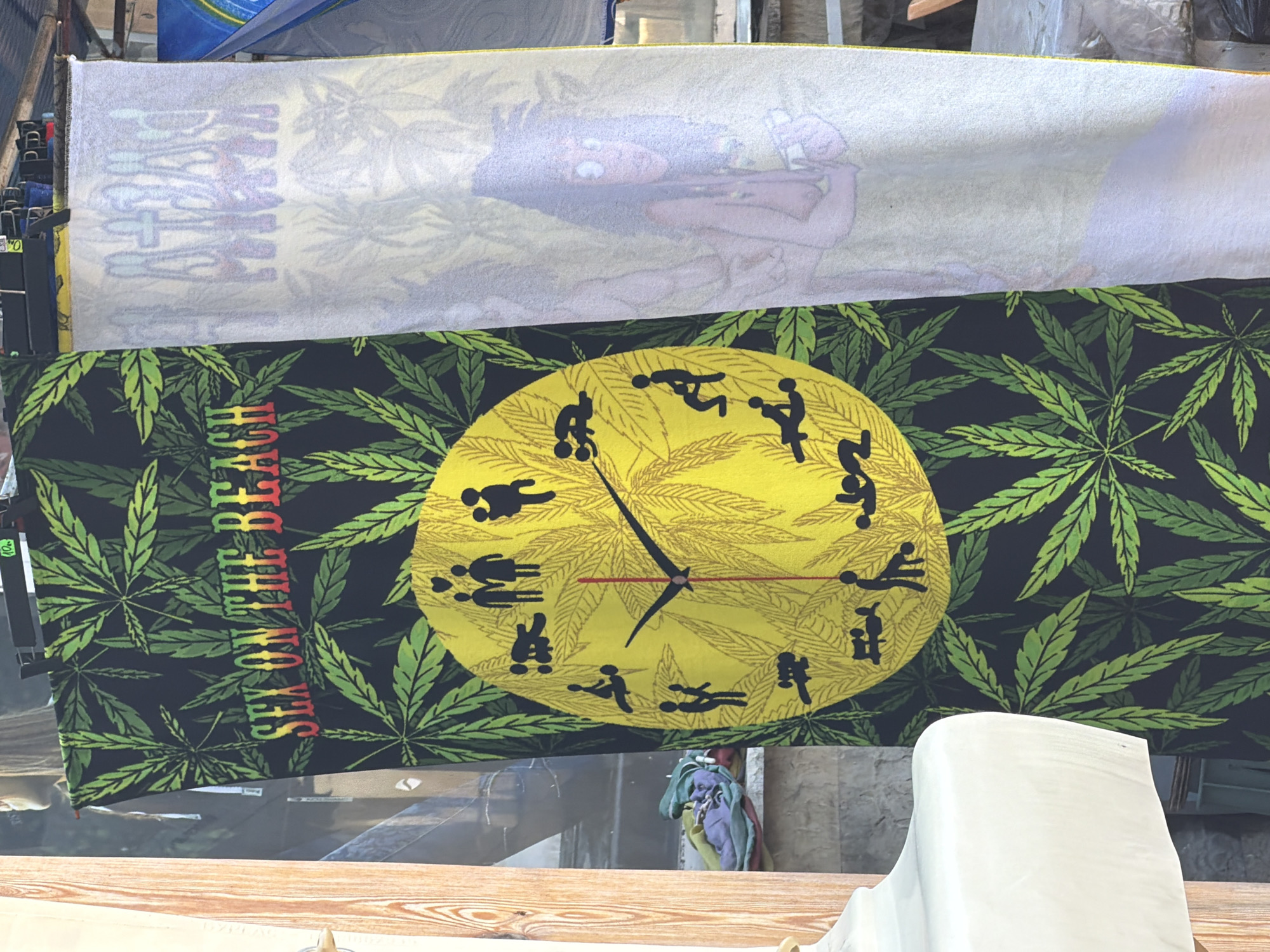 The image depicts a street scene in Tsarevo, Bulgaria, with two banners hanging side-by-side. The left banner features a black background with green cannabis leaves and the red, yellow, and green text "SEX ON THE BEACH". In the center of this banner is a large yellow clock face, also decorated with cannabis leaves, and marked with silhouettes of various human figures engaged in sexual acts. The right banner is a white fabric with a faint image of a cartoonish woman with dark curly hair and large eyes. The banners are suspended from a metal structure, likely a stall or display. In the background, a wooden plank is visible on the left, and various objects and structures suggest an outdoor market or vendor setting.