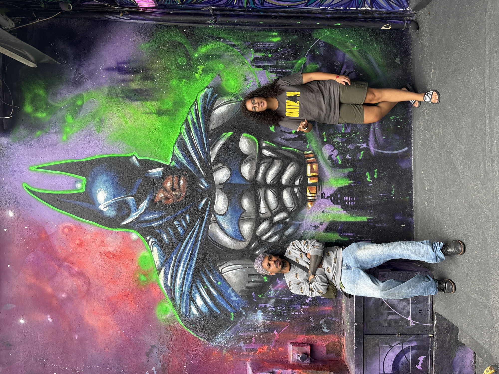 Two people are posed in front of a mural of Batman. One person is lying on the left side of the image in blue jeans, a gray shirt with Batman symbols on it, and black shoes. They have short, dyed hair and tattoos on their arms. The second person is lying on the right side of the image. They are wearing a gray t-shirt with the word “Batman” on it in yellow, olive green shorts, and black and white sandals. They have curly hair and glasses. The mural is an illustration of Batman with a city skyline in the background. The colors of the mural include purple, green, red, blue, gray, and black. A small Batman logo is on a gray door in the lower right corner. Based on available information, this location is in the city of São Paulo, Brazil.