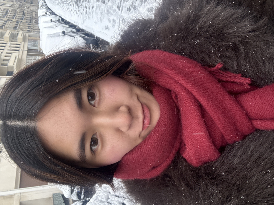 A female subject, appearing to be of East Asian ethnicity, is depicted in a close-up, outdoor portrait shot during a snowfall in Paris, France. She has dark brown hair parted in the middle, brown eyes, and a slight, closed-lip smile. Her cheeks show a faint flush. She wears a dark brown, possibly faux fur, coat and a thick, cranberry-red woven scarf wrapped around her neck. Small snowflakes are visible on her hair, scarf, and coat, indicating active precipitation.
The background reveals a street scene typical of Paris. A row of parked vehicles, covered in a fresh layer of snow, lines the right side of the frame. The ground, likely a paved street or cobblestones, is also covered in snow, showing some disturbed areas or tracks. Multi-story buildings with light-colored facades, numerous windows, and classic Parisian architectural elements, including balconies, are visible further down the street, confirming the urban location in Paris. The lighting is soft and diffused, consistent with an overcast, snowy day.