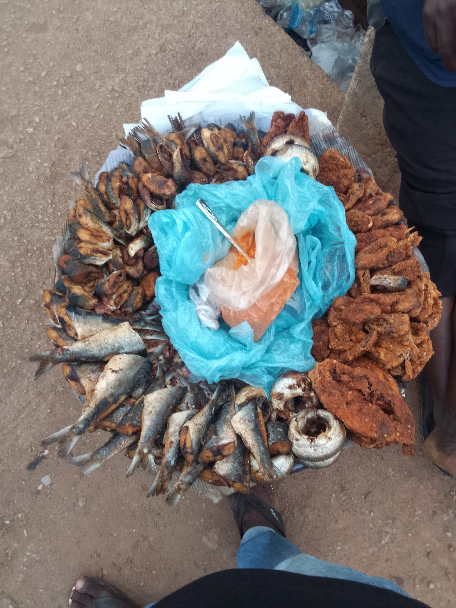 A high-angle medium shot shows an array of fried fish and other fried items displayed on a round tray. The tray is lined with newspaper and white paper towels, and a bright blue plastic bag containing what appears to be spices is placed in the center. The fried fish are arranged around the bag. To the right of the fish are pieces of what appear to be fried yam. A person is standing to the right of the tray, wearing black pants and tan sandals. Another person, wearing jeans, is visible in the lower center of the image. The location is reportedly Bukuru, Nigeria. The surface beneath the tray appears to be unpaved ground.