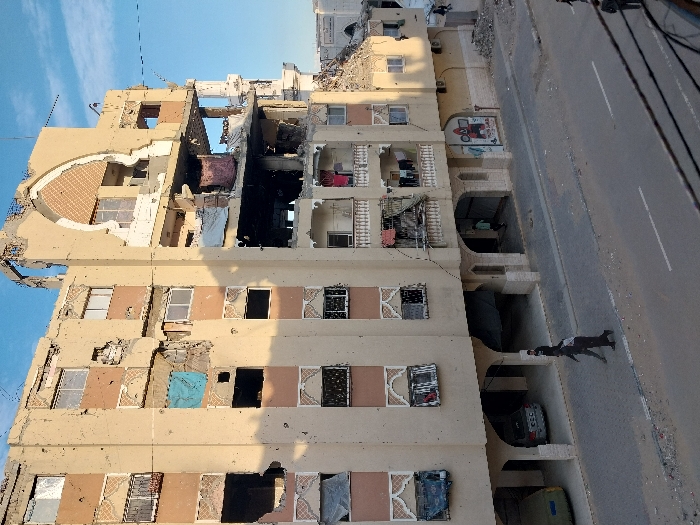 The image depicts a multi-story building that has suffered extensive damage, likely from an explosion or conflict. The building, constructed with a light beige or sandy-colored facade, dominates the left side of the frame and stands alongside a paved road.
Severe structural damage is evident, particularly on the upper floors, where large sections of exterior walls and internal partitions are completely missing, exposing concrete slabs and rebar. A prominent area in the mid-to-upper section of the building is heavily charred and blackened, indicating significant fire damage. Through the destroyed sections, glimpses of interiors, including a dark pink or red piece of fabric hanging from a damaged balcony, are visible. Many windows are shattered, missing, or have warped frames. The building's facade is also marked with numerous smaller holes, consistent with shrapnel or projectile impacts.
Architectural features include decorative patterns around some windows on the lower floors and large arched openings at ground level. A distinctive large, semi-circular recessed area with patterned tiling is visible on the upper left side of the structure. Laundry appears to be hanging on some of the surviving balconies.
At the ground level, under the arched sections, two cars are partially visible, parked, along with a green container, possibly a dumpster. On the wall beneath one of these arches, a sign or mural displays a red tractor with a plow, accompanied by indistinct Arabic text above it.
Adjacent to the damaged building is a paved road featuring a white dashed center line, indicating two lanes. Scattered debris is visible on the road surface. Utility lines are stretched across the scene, presumably connected to poles or structures outside the frame.
A single person, dressed in dark clothing, is walking on the road or sidewalk in the lower right portion of the image, moving away from the viewer.
The scene is illuminated by natural daylight under a clear blue sky with a few faint clouds, suggesting either morning or afternoon. The overall impression is one of significant destruction in an urban or residential environment.
