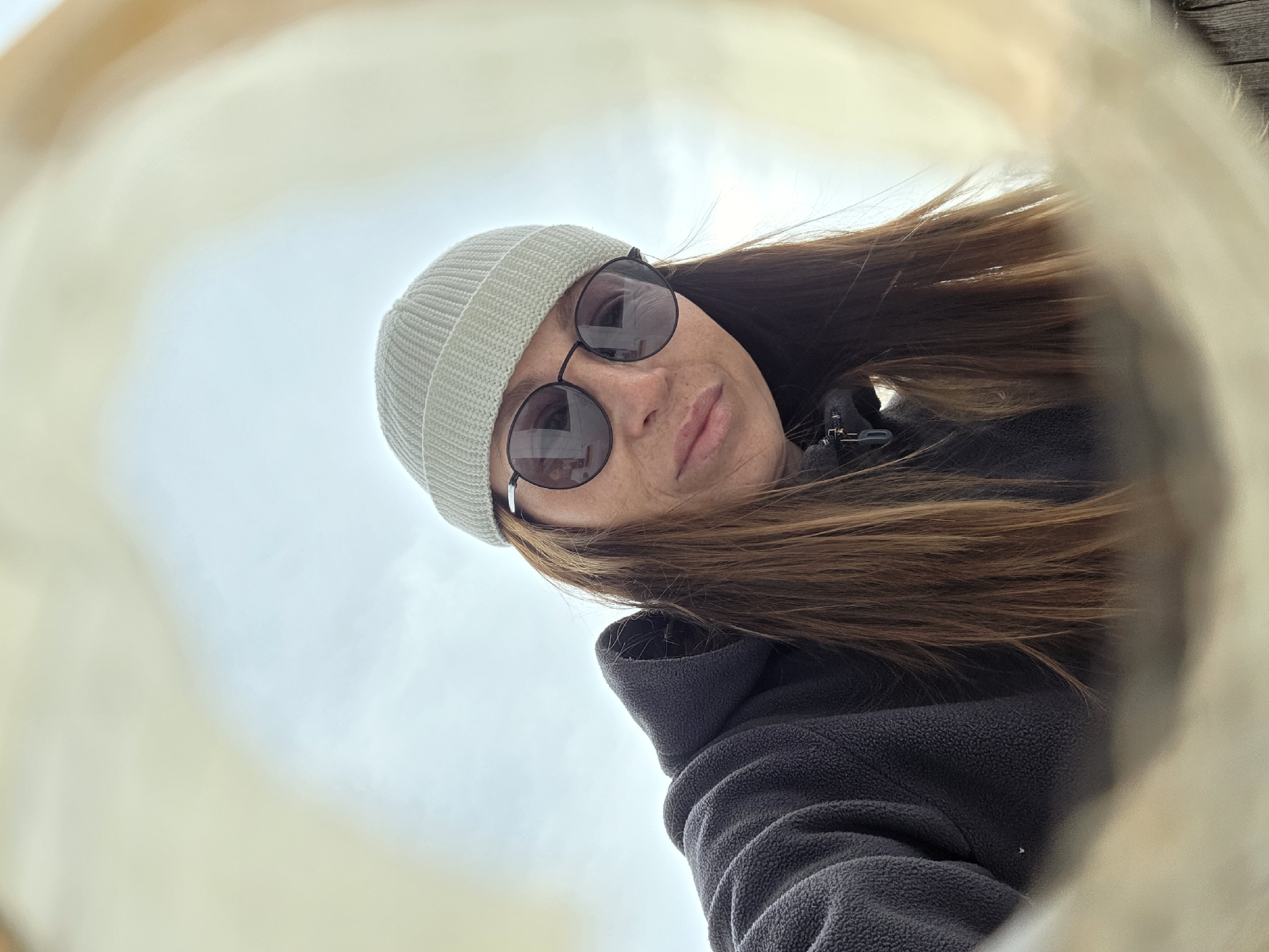 A person is looking down into a round opening, wearing a light gray knit hat, round sunglasses, a dark fleece jacket, and has long brown hair. The background visible through the opening appears to be a light gray sky. The location is Unknown, Unknown.