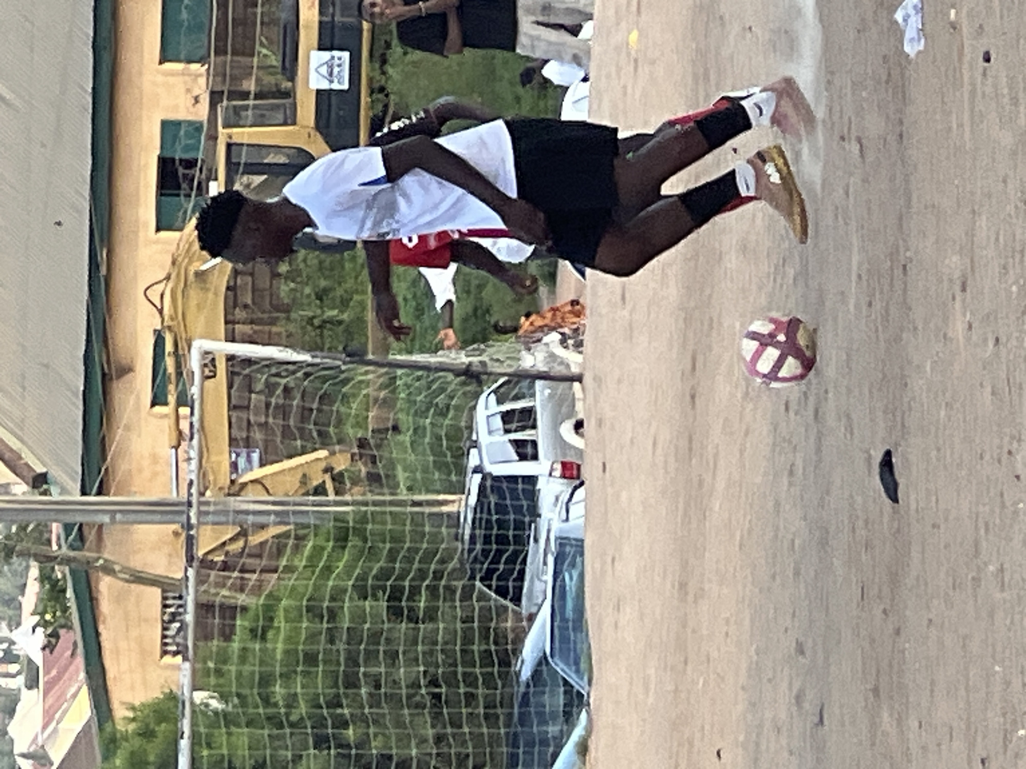 The image shows a group of young men playing soccer on a dirt field. In the background, there is a soccer net, a yellow construction vehicle, several buildings with corrugated metal roofs, and two parked cars. One car is a silver SUV, and the other is partially submerged in what appears to be sand or mud. The sky is overcast. The overall impression is of an informal soccer game taking place in a developing urban or semi-urban environment. The scene is likely in Jos, Nigeria, as indicated by the specific request.
